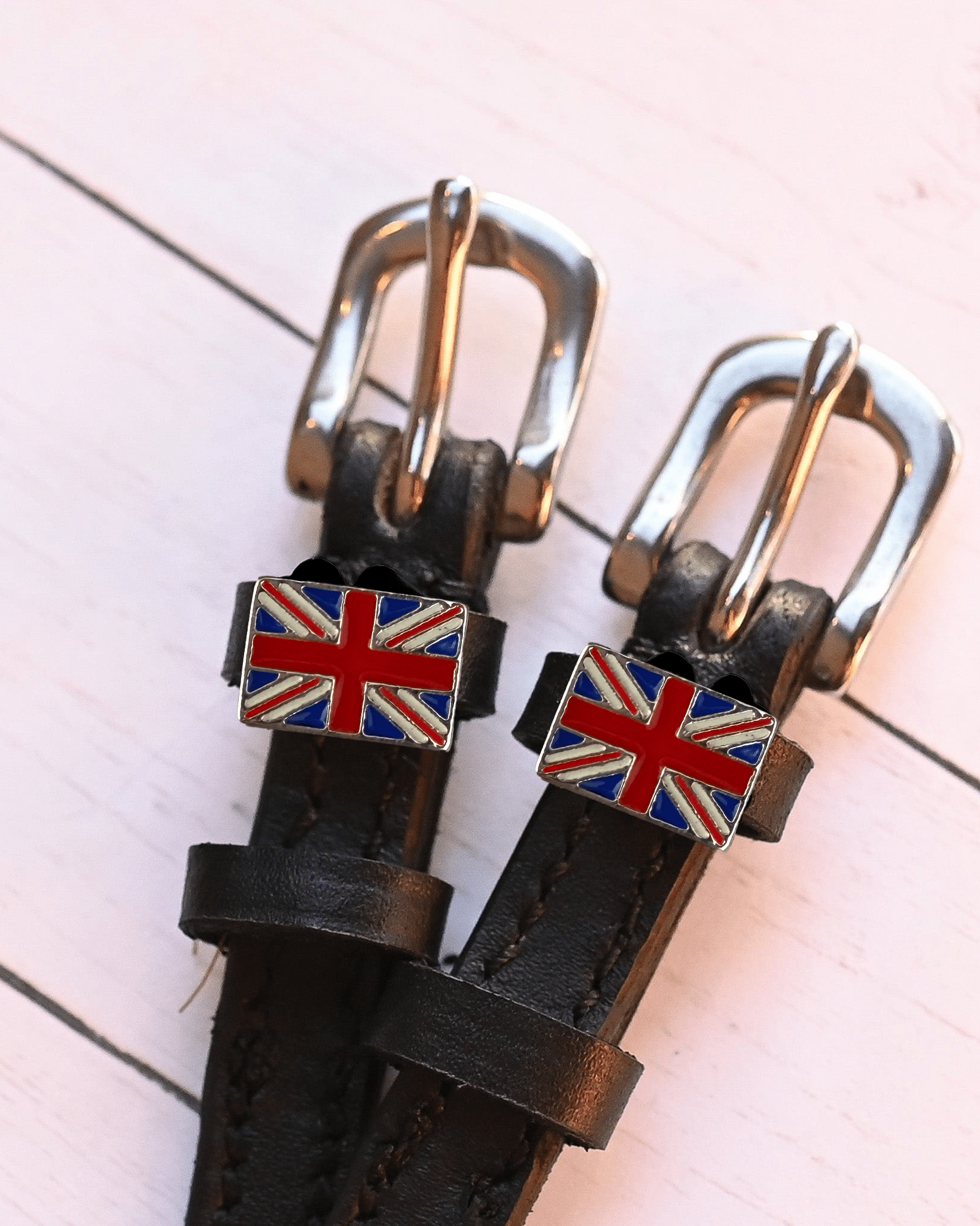 Overhead of black leather spur straps with silver buckles and Union Jack charms on pink wood