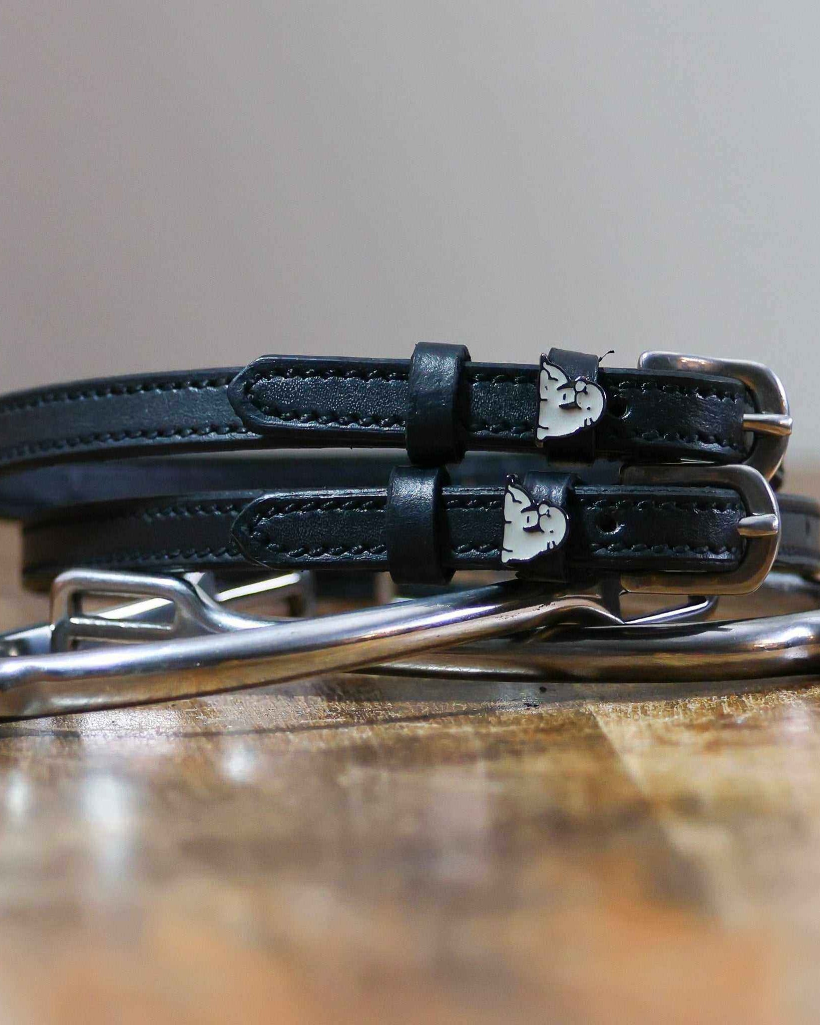 Black leather walrus spur straps with silver buckles angled on white wooden planks