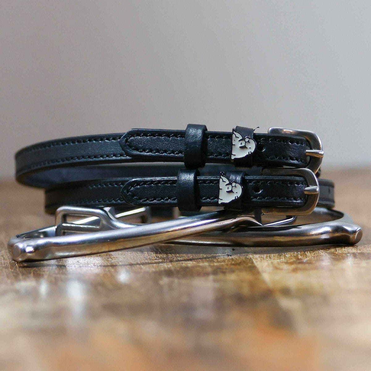 Black leather walrus spur straps with silver buckles angled on white wooden planks