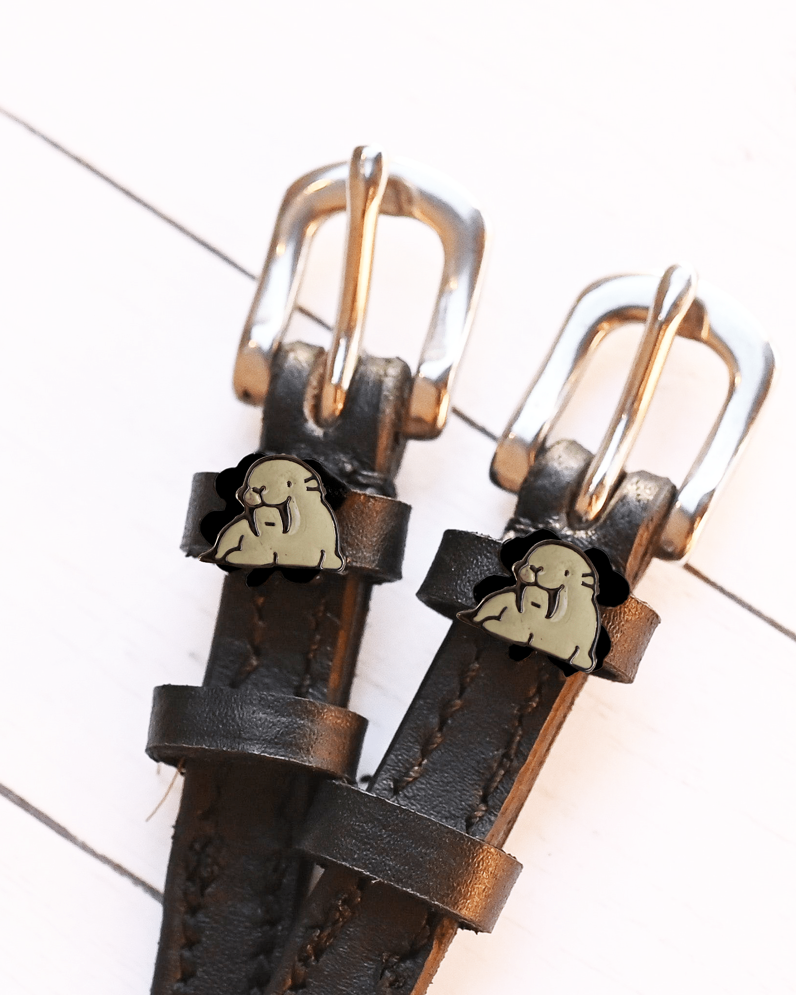 Grey walrus charm spur straps with silver buckles displayed on white wood planks