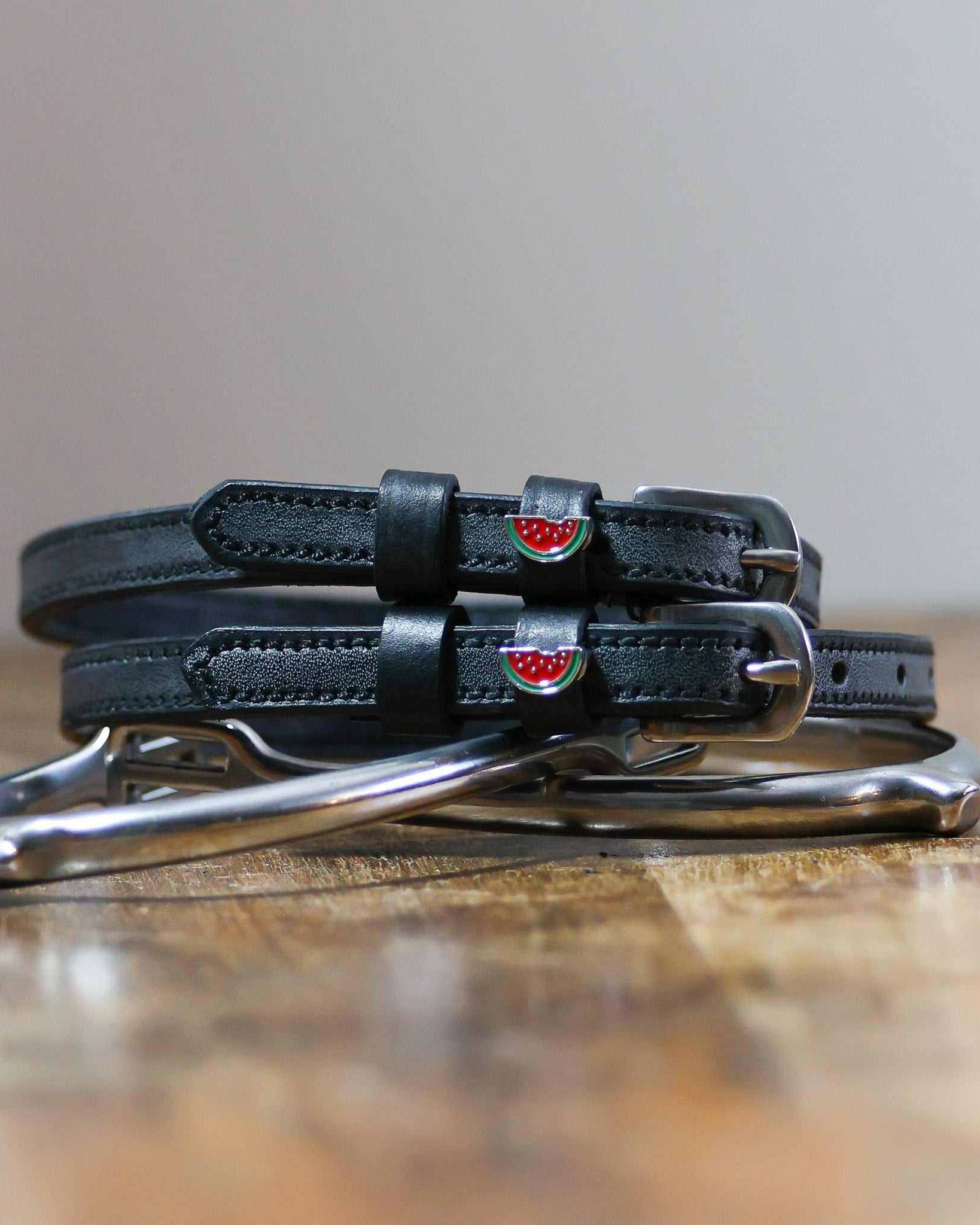 Black watermelon charm spur straps coiled beside a silver spur on a rustic wood surface