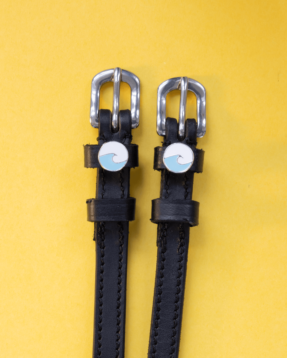 Blue wave charm spur straps in black leather flat-laid on a yellow background