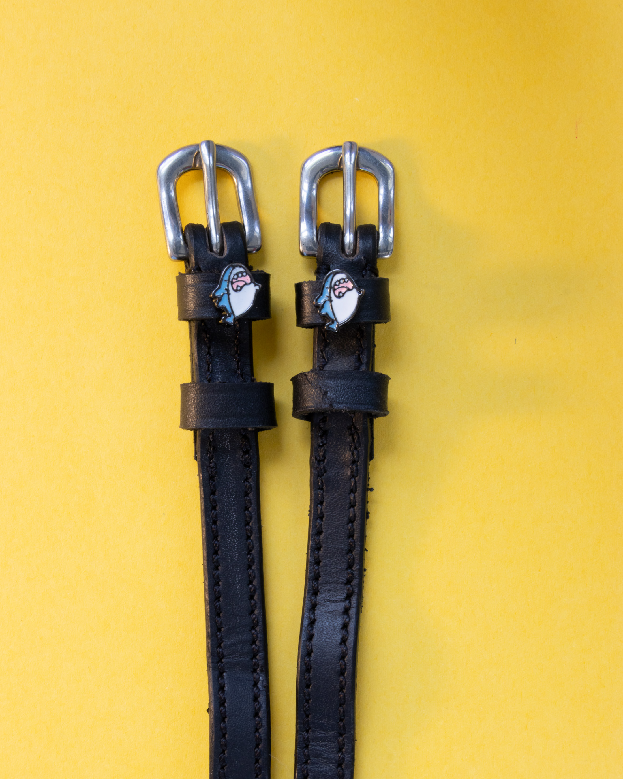 Pair of black leather shark charm spur straps with silver buckles on a yellow background
