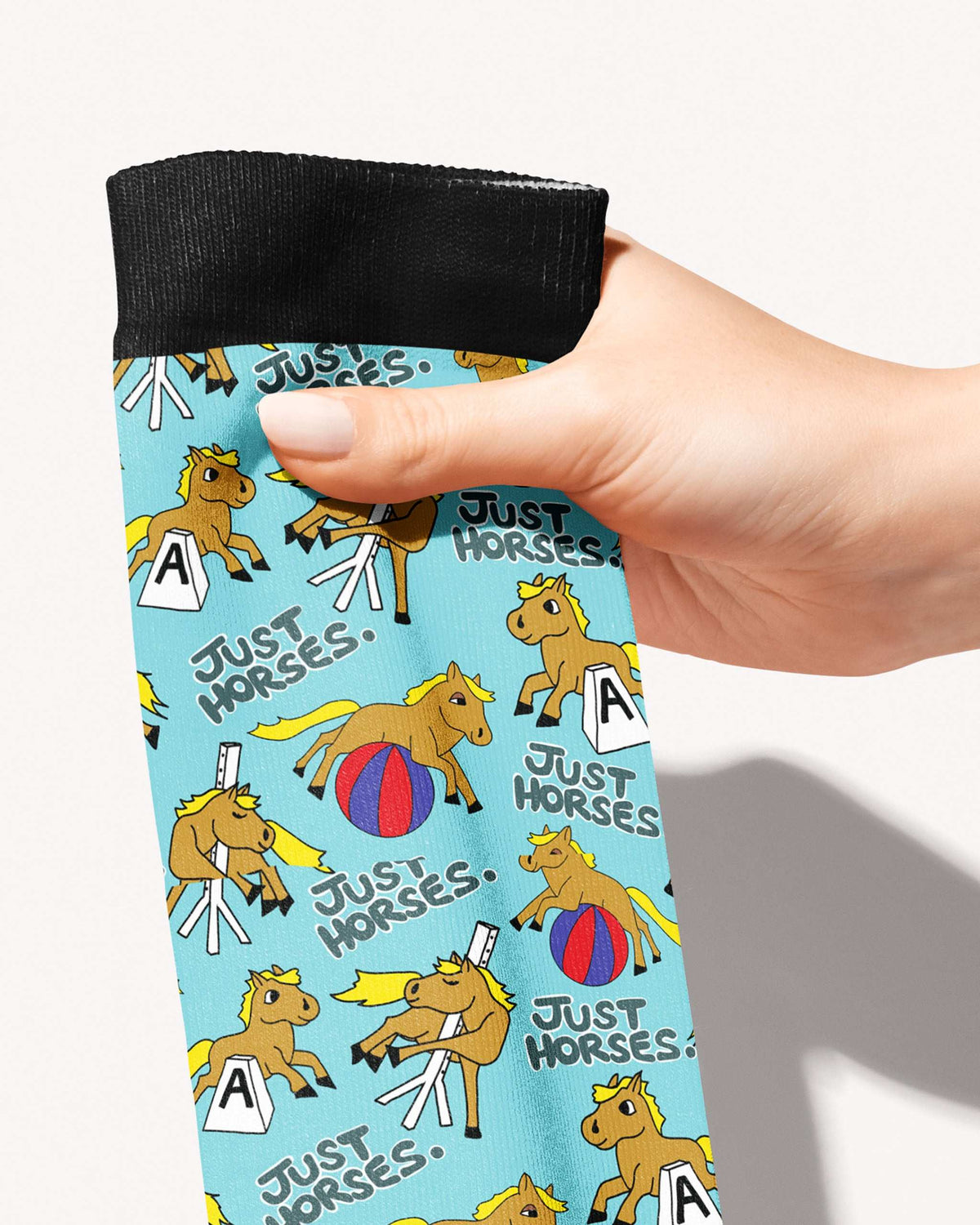 A close-up of a hand holding a light blue patterned sock with a black ribbed cuff. The sock features a repeating cartoon horse design with the text 'JUST HORSES.