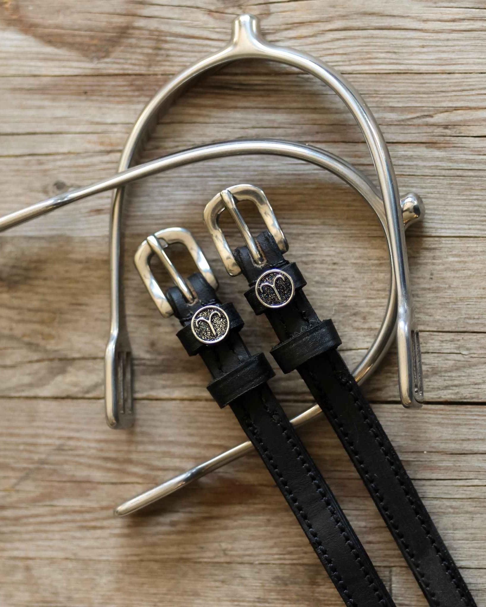 Black leather Aries Spur Straps with stainless steel buckles and custom Aries charm on wooden background
