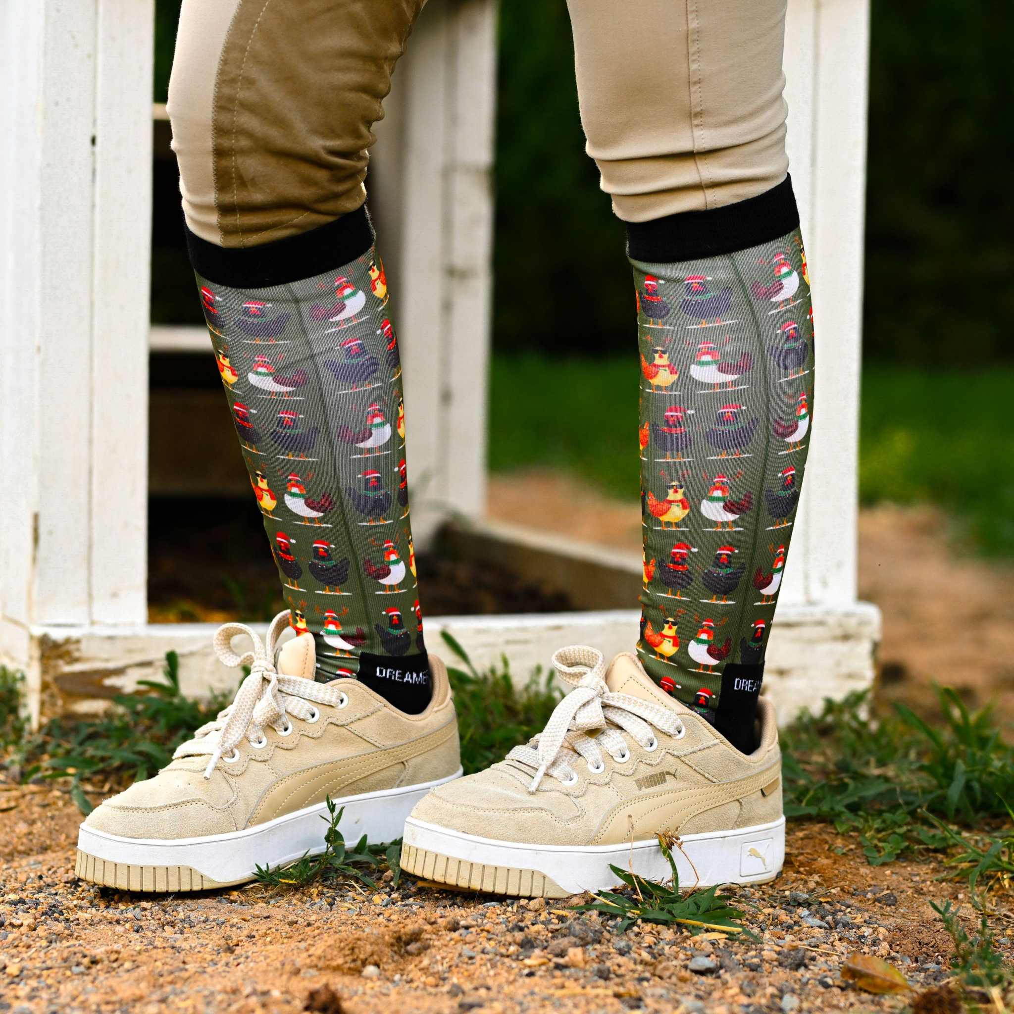 Holiday-themed boot socks with colorful chicken pattern and black stretchy top, worn with beige pants and cream sneakers outdoors.
