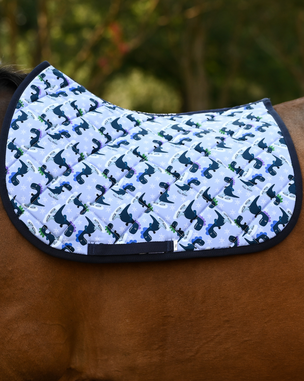 Dino-skate saddle pad in pale periwinkle with navy skating dinosaurs, shown on a bay horse without a saddle