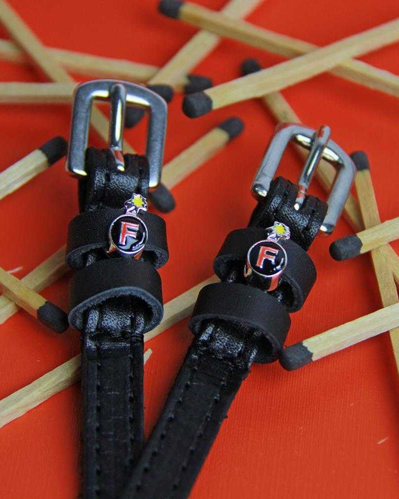 Close-up of PlainJane black leather spur straps with silver buckles from the Ultimate F-Bomb Pack on orange background with scattered matchsticks