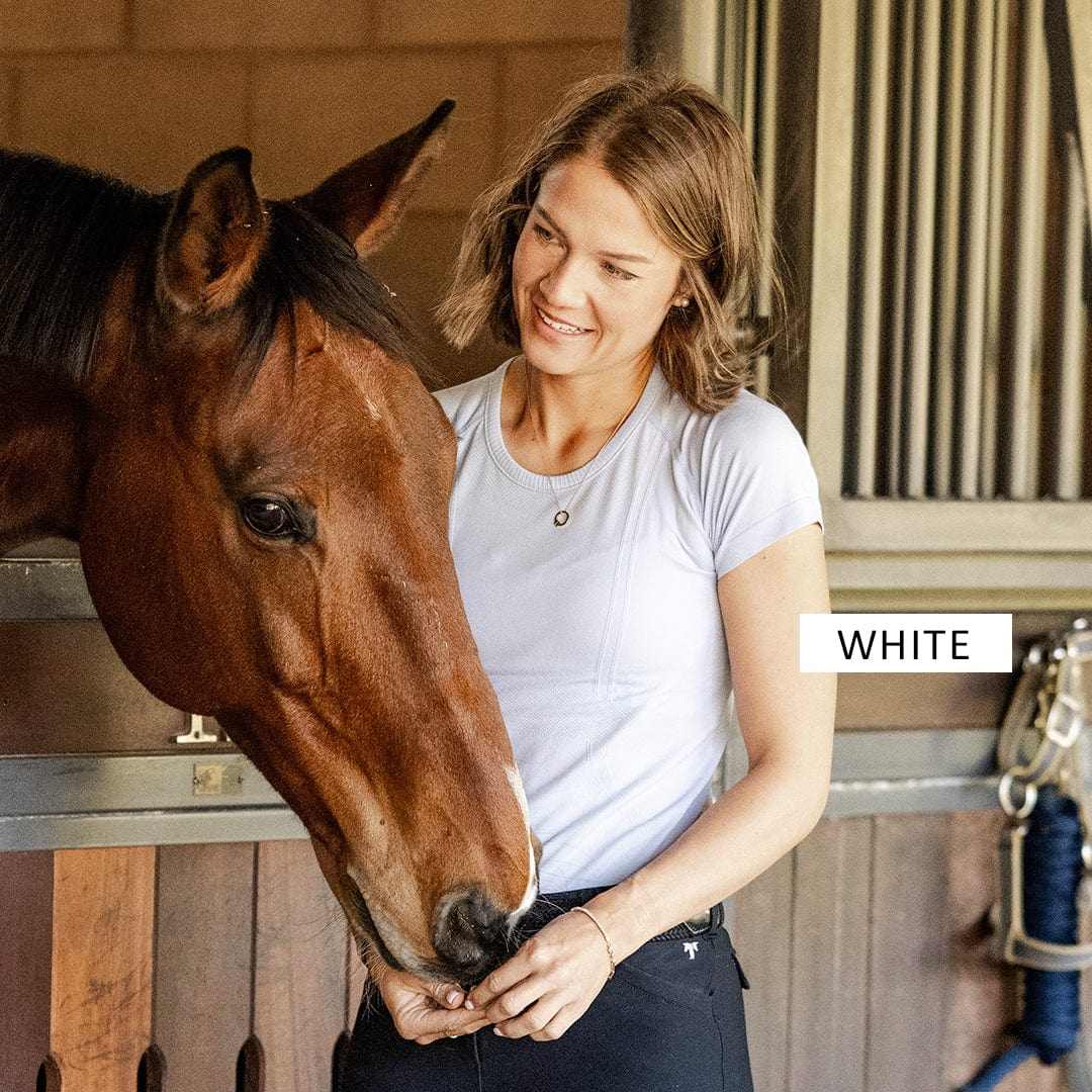Woman wearing white Final Sale Equitation Tech Short Sleeve Top standing next to a brown horse in a stable active wear
