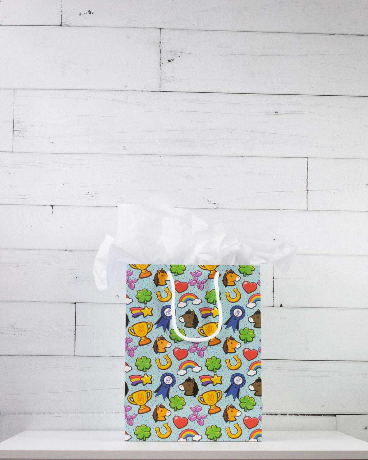 Full O' Luck medium gift bag on a white shelf, bright print of trophies, rosettes and four-leaf clovers.