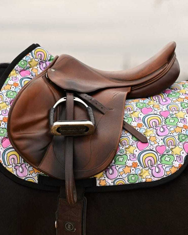 Brown leather saddle on horse with colorful Lucky Equestrian Bundle Full O’ Luck saddle pad featuring clovers, rainbows, and horseshoes