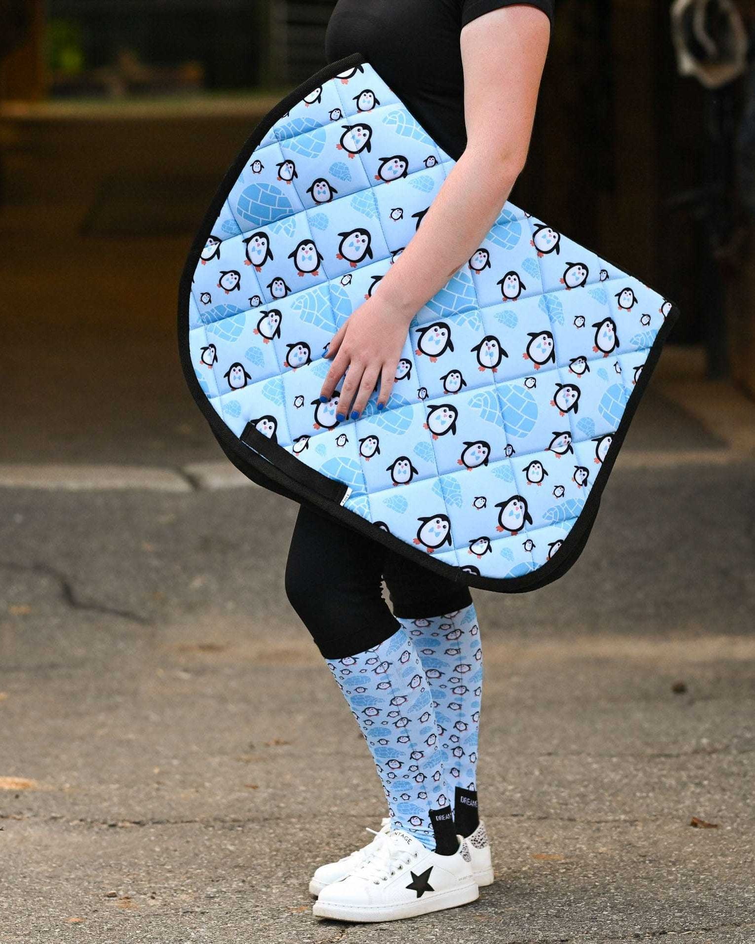 Person holding blue Igloo Crew Saddle Pad with penguin pattern, matching socks, outdoor barn aisle background