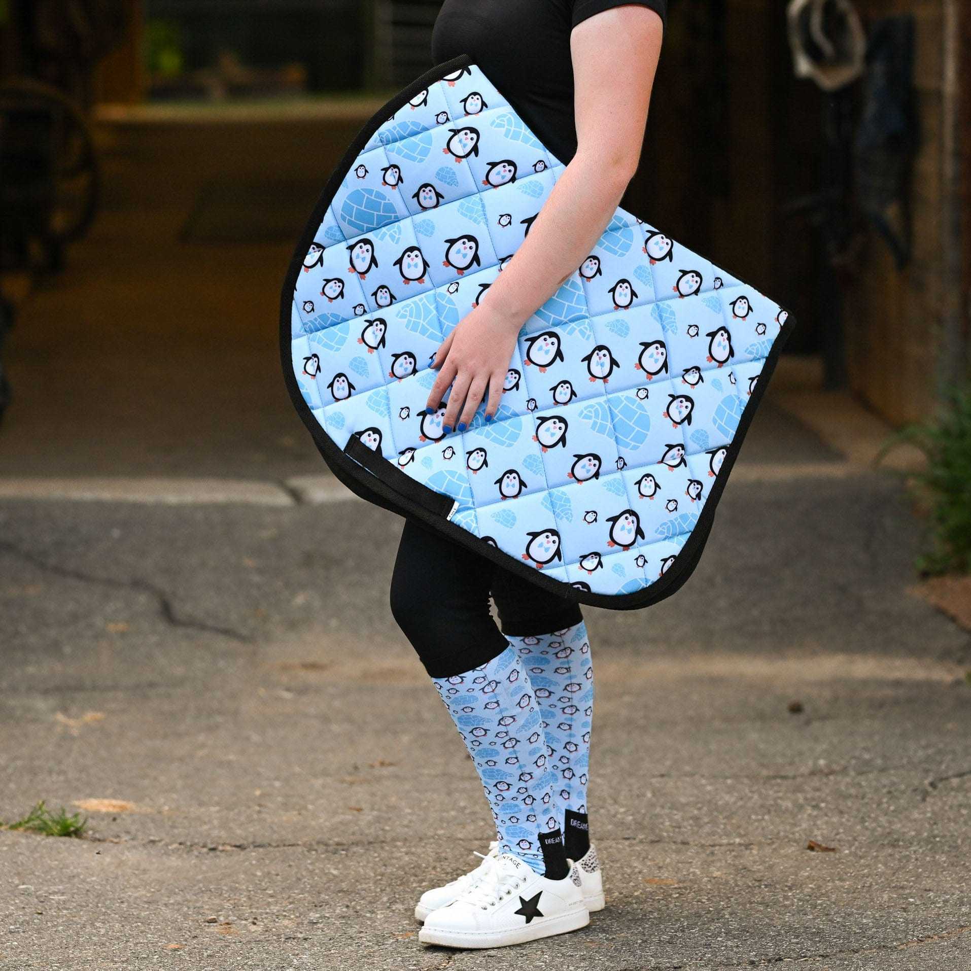 Person holding blue Igloo Crew Saddle Pad with penguin pattern, matching socks, outdoor barn aisle background