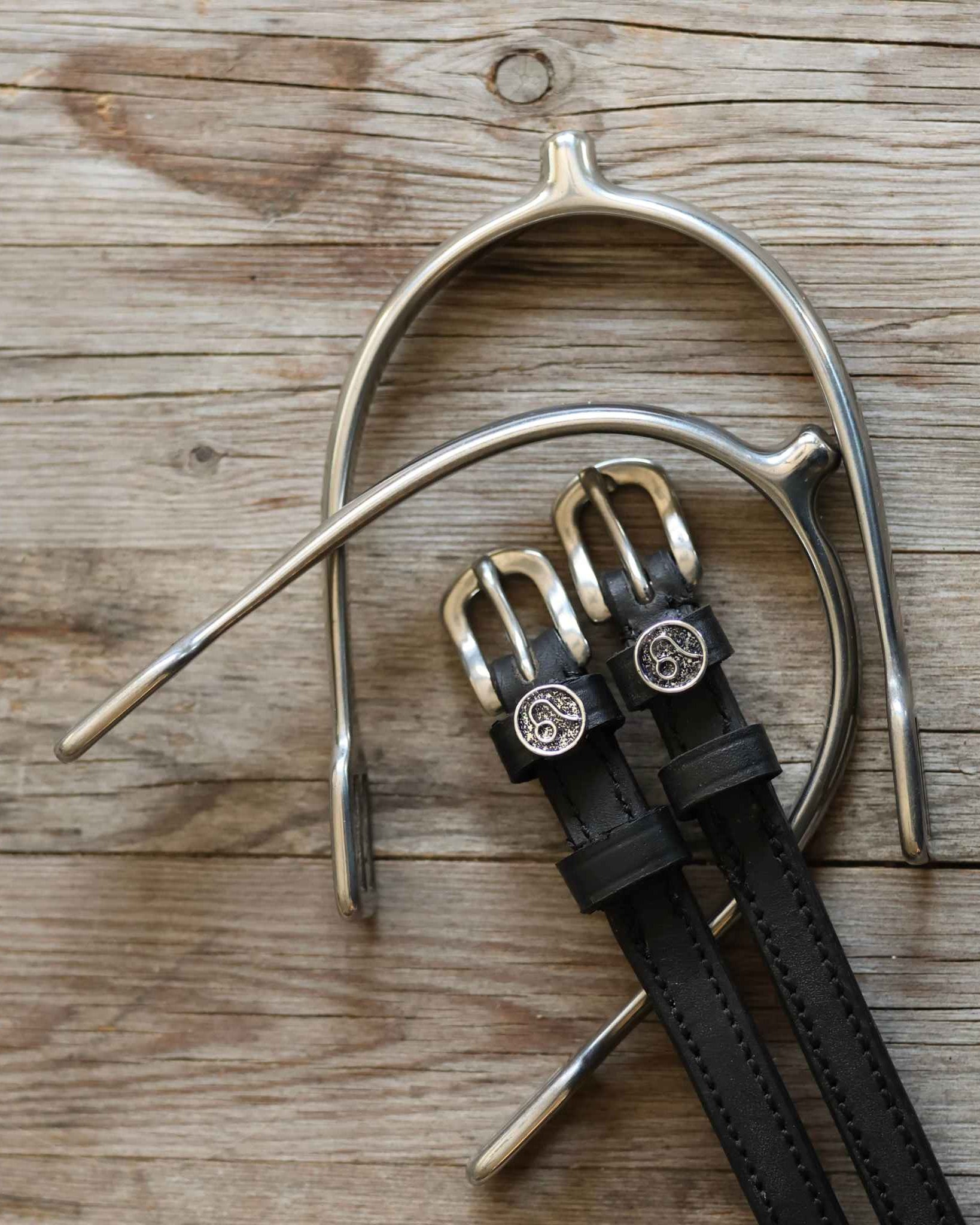 Black leather Leo Spur Straps with stainless steel buckles and custom ManeJane charms on wooden background