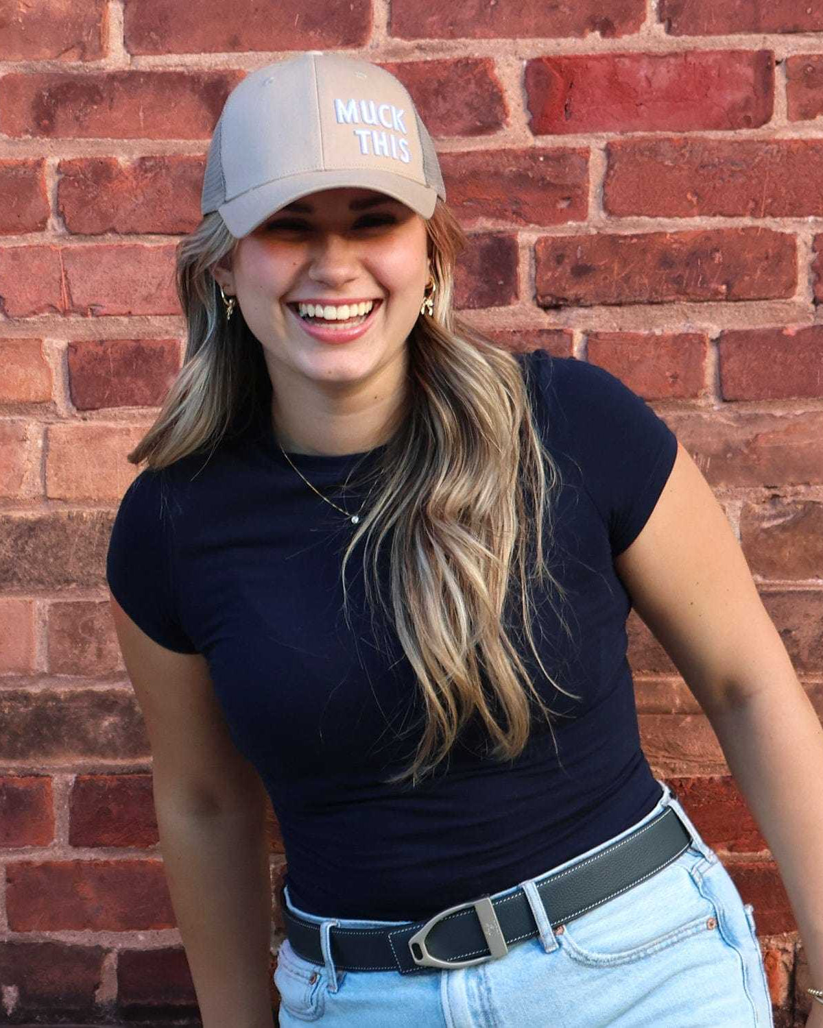Woman wearing Muck This trucker hat, navy shirt, and jeans against brick wall