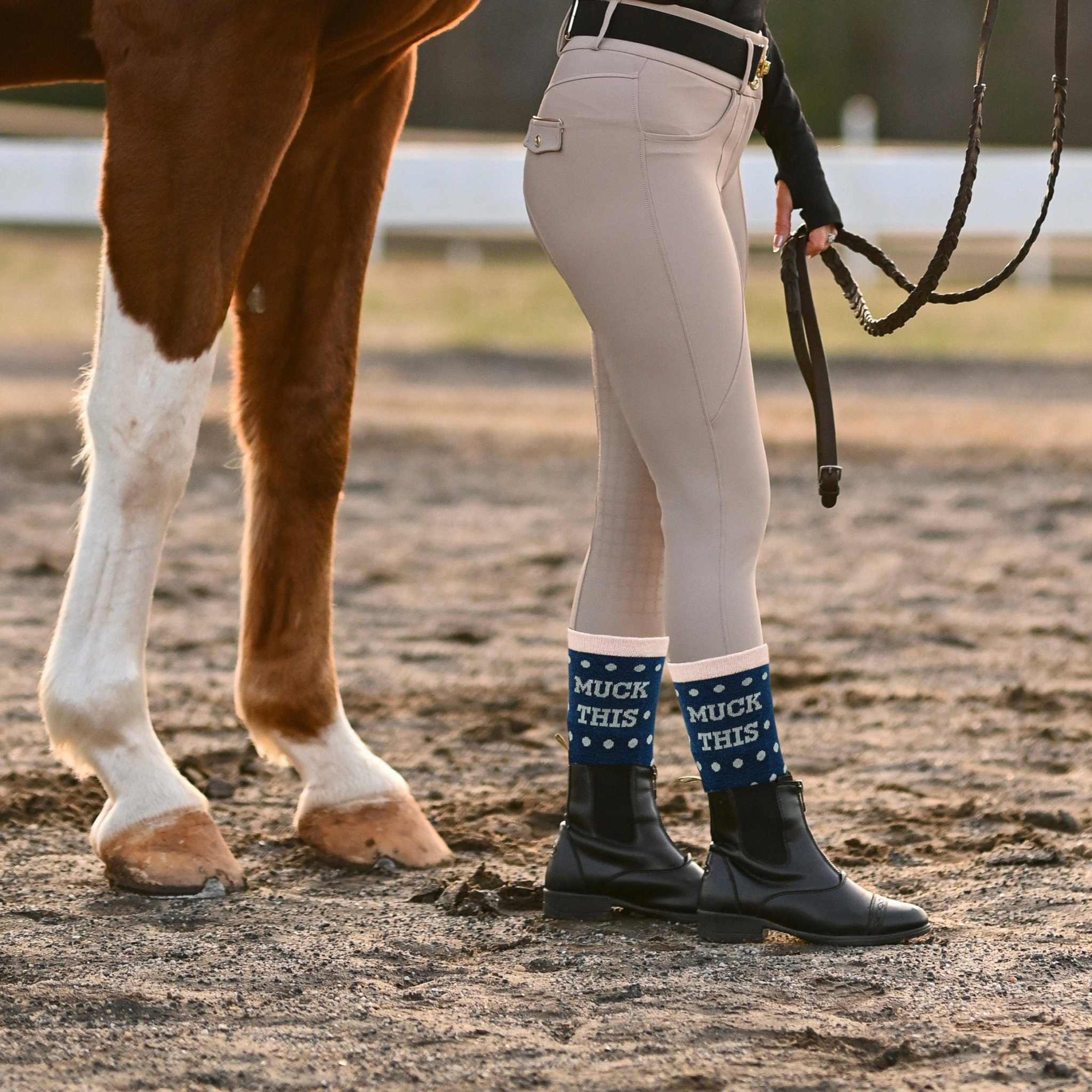 Equestrian wearing "Muck This" socks and boots standing next to a horse outdoors