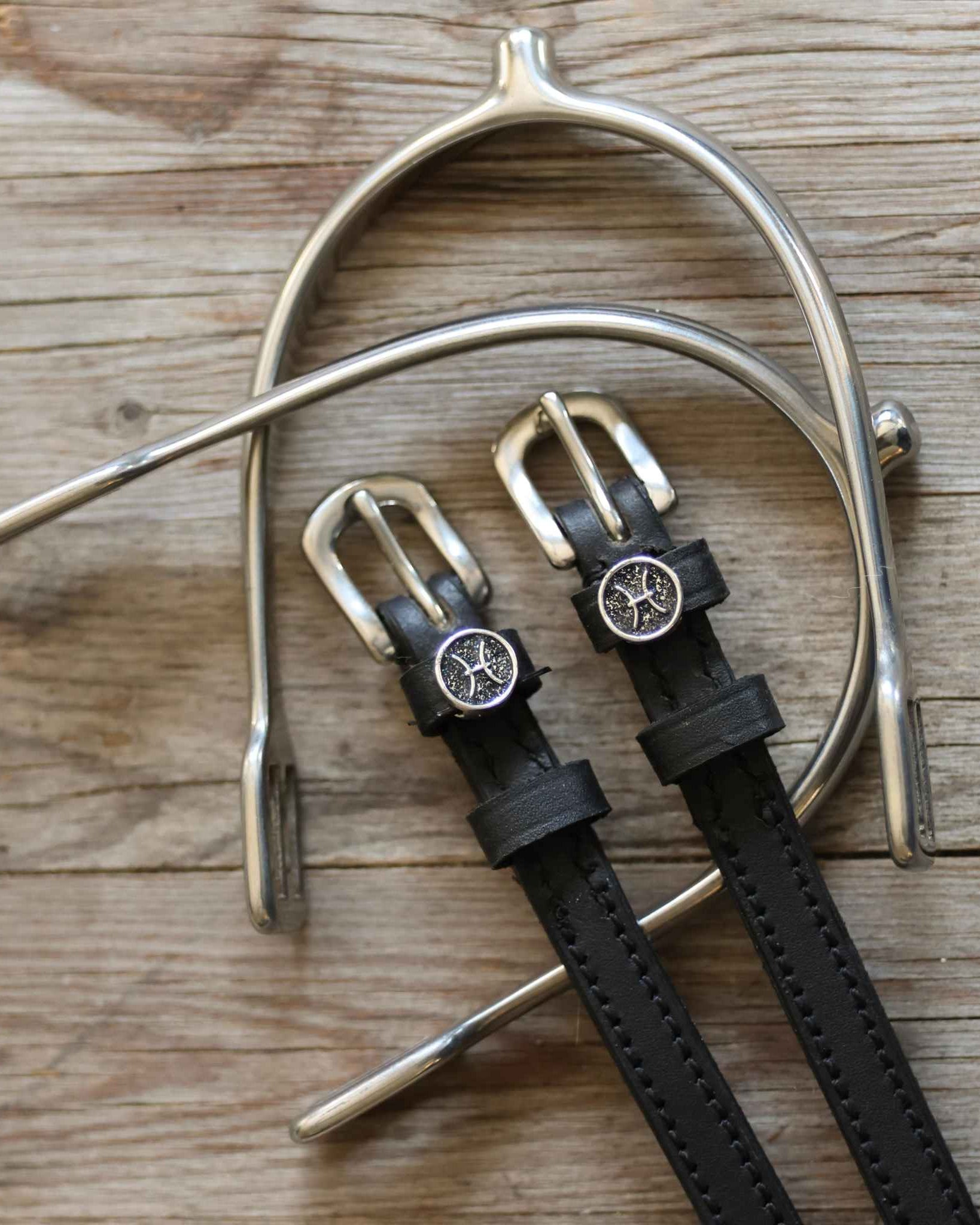 Pisces Spur Straps black leather with stainless steel buckles and custom charm on wooden background