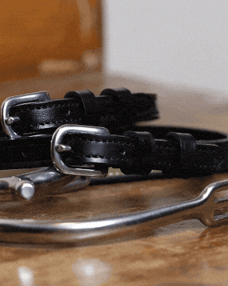 Plain black leather ManeJane spur straps with no charm, coiled by a stirrup on wood