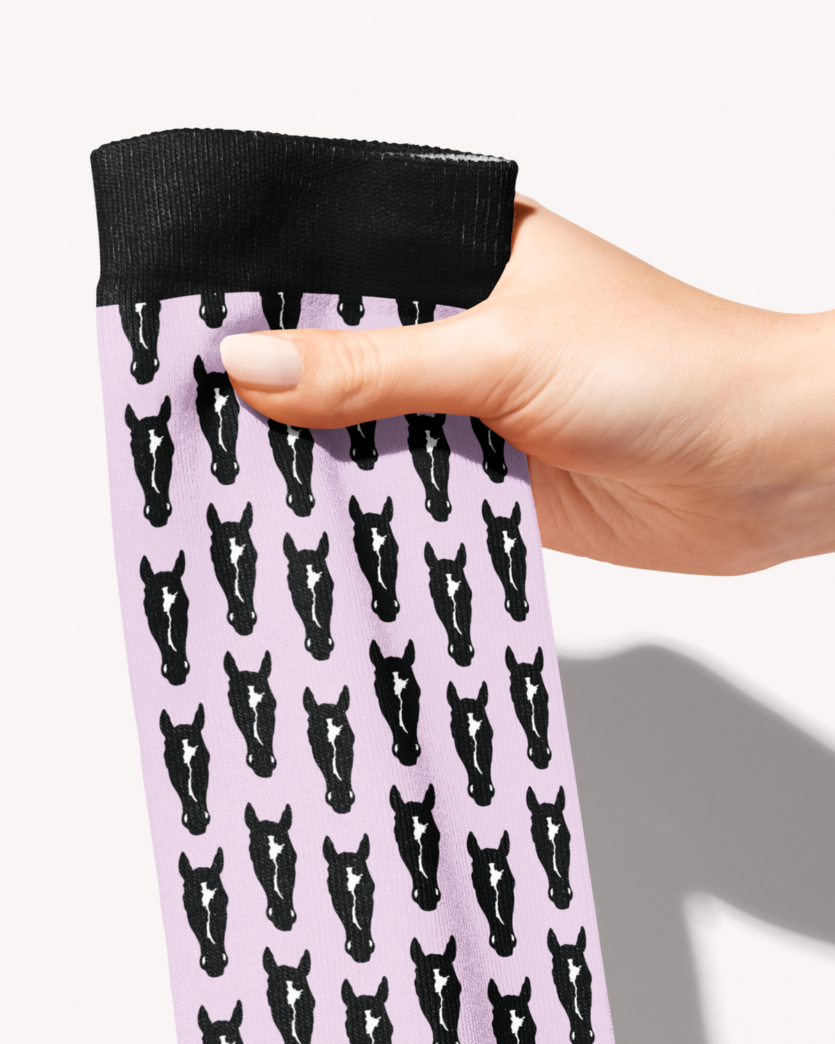 Hand holding the black cuff of the Nikko boot sock, close-up of black horse heads with white blazes on pale pink