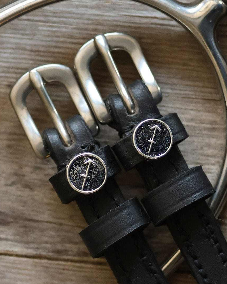 Black leather Sagittarius Spur Straps with stainless steel buckles and custom charm on wooden surface