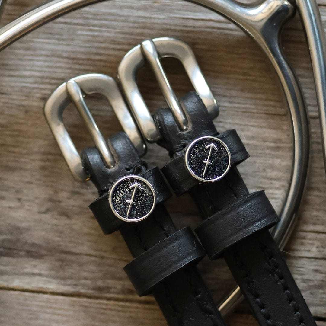 Black leather Sagittarius Spur Straps with stainless steel buckles and custom charm on wooden surface
