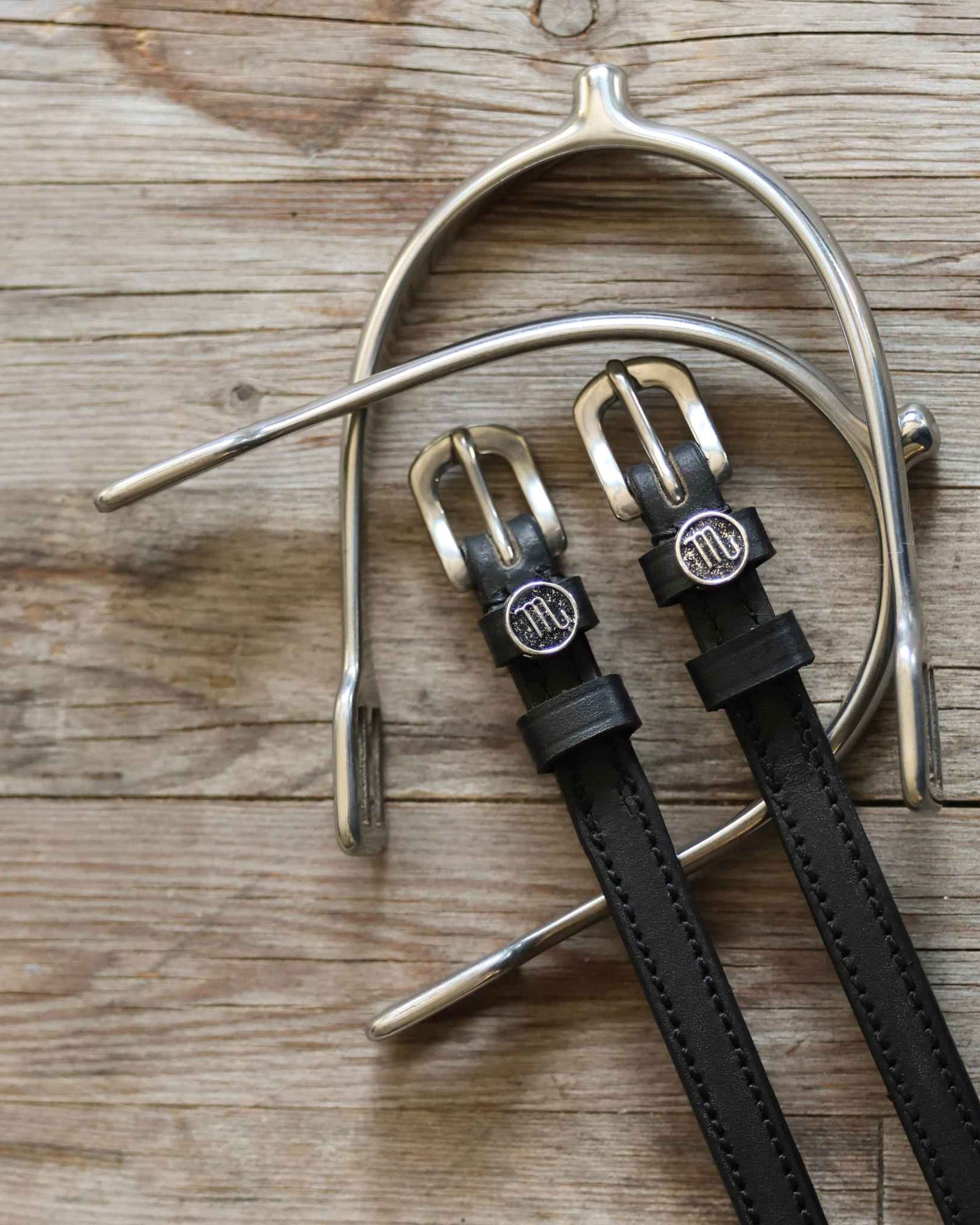 Black leather Scorpio Spur Straps with stainless steel buckles and custom charm on wood background