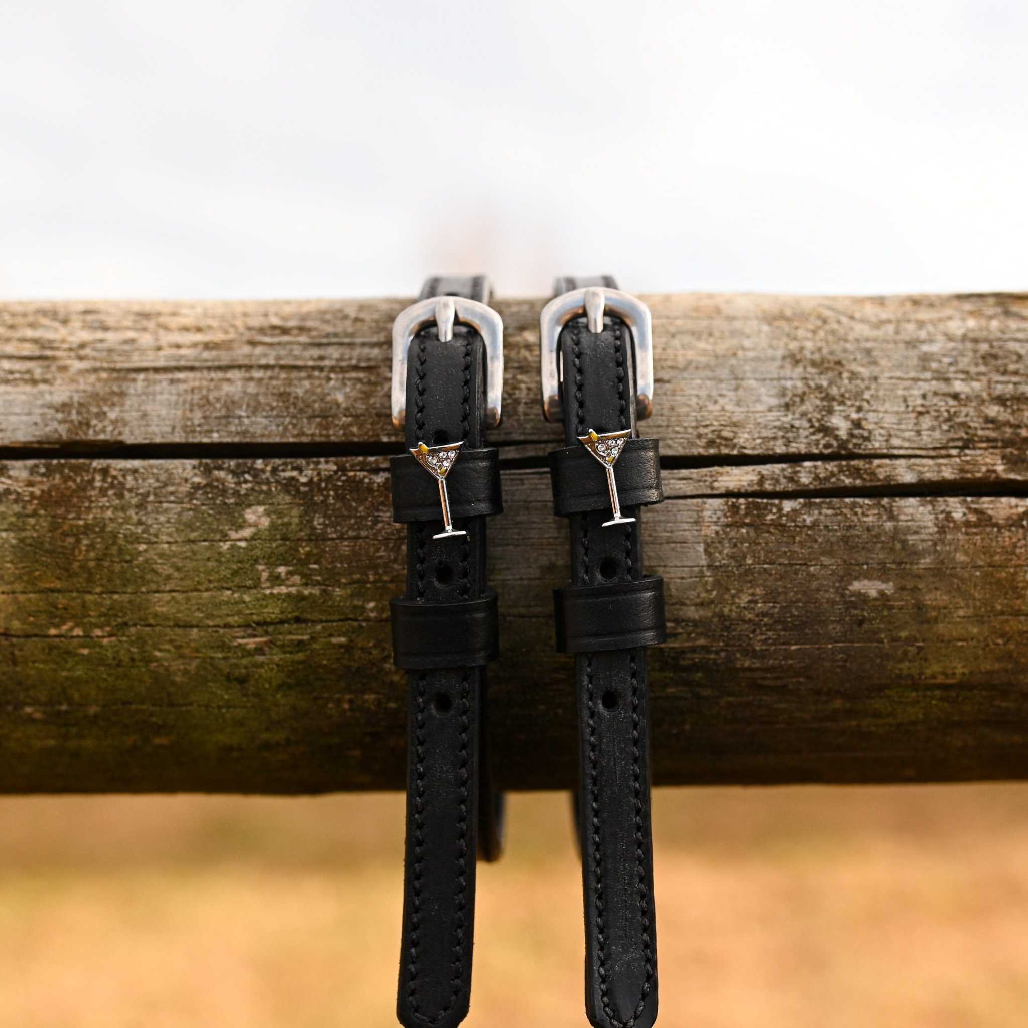 Black leather spur straps with silver martini glass charm and stainless steel buckles hanging on wooden beam