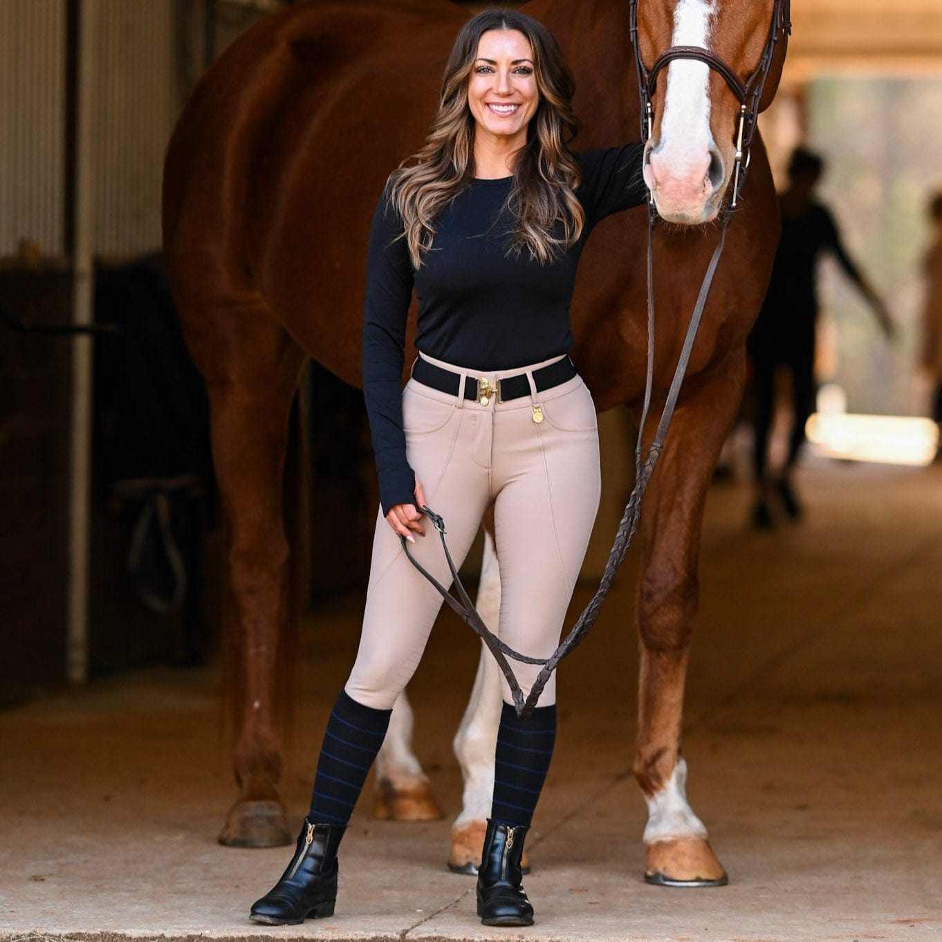 woman wearing knit boot socks with riding boots standing next to a horse in stable