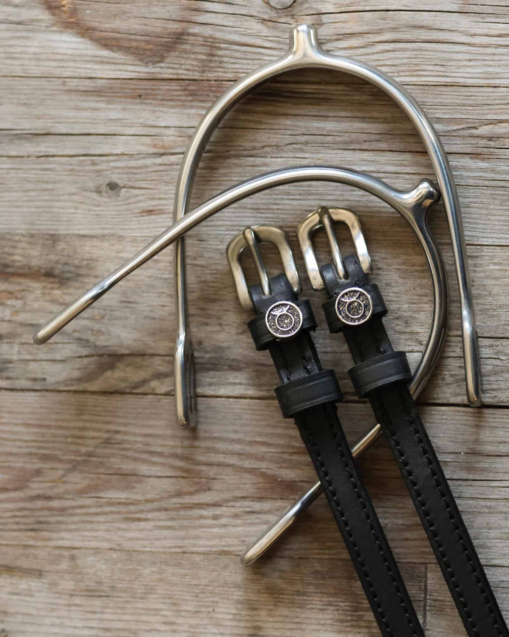 Taurus Spur Straps black leather with stainless steel buckles and custom charms on wooden surface with metal spurs