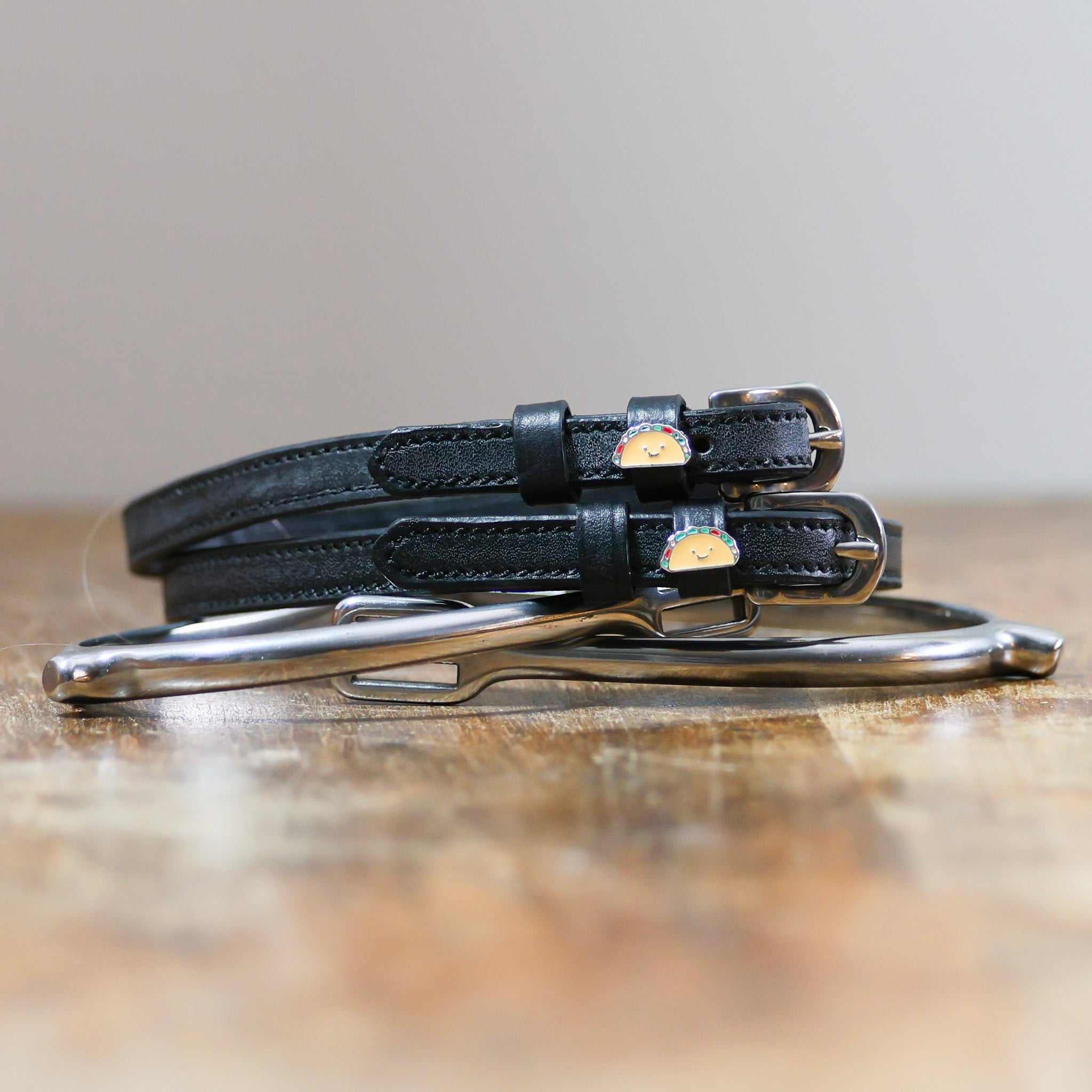 Black leather spur straps with stainless steel buckles and custom charms on wooden surface