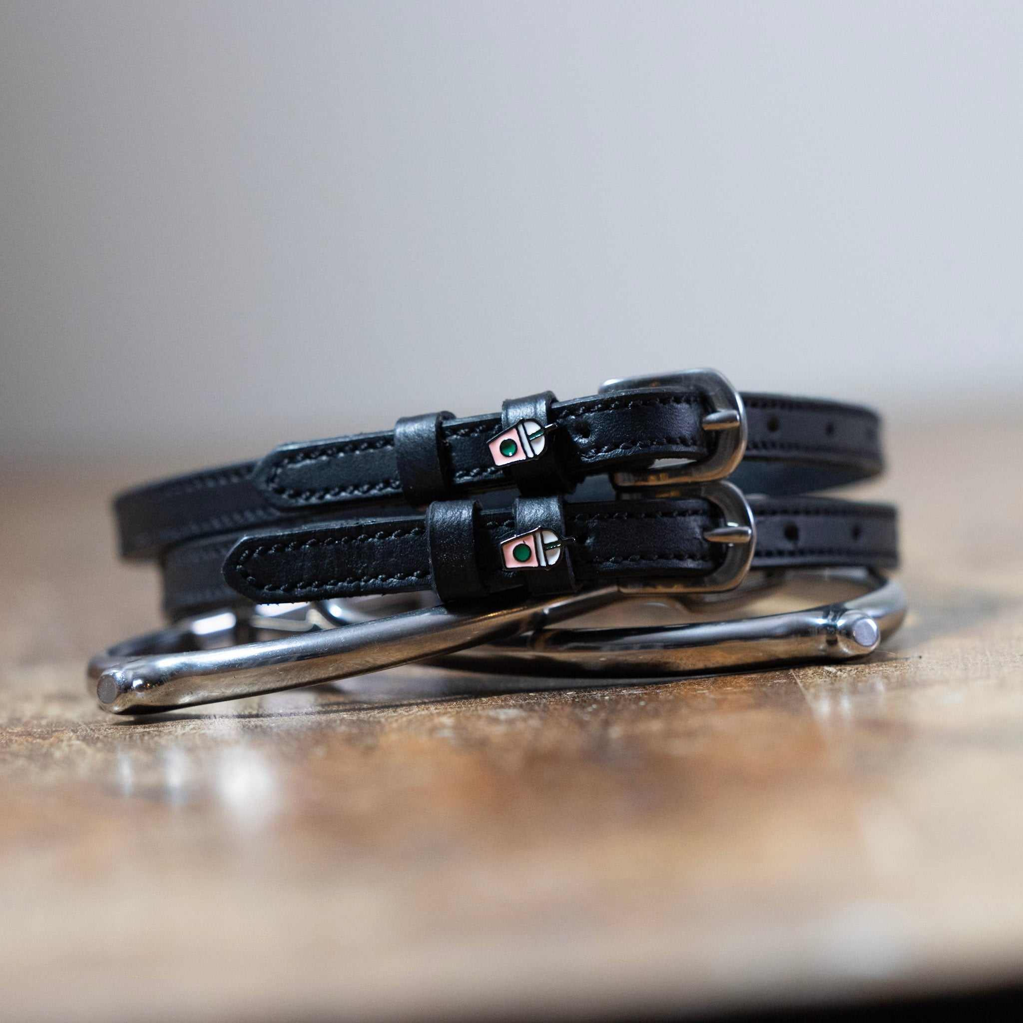 Black leather Frappe Spur Straps with stainless steel buckles and custom charm on wooden surface