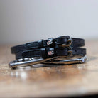 Black leather Frappe Spur Straps with stainless steel buckles and custom charm on wooden surface