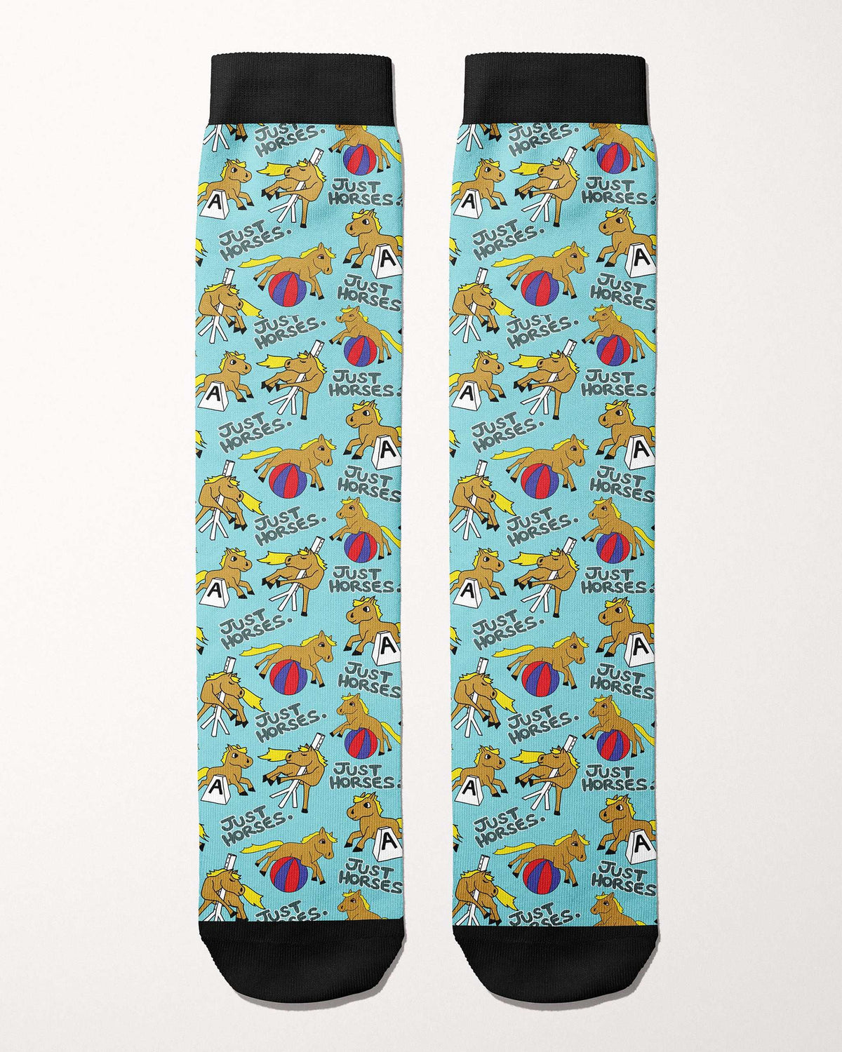A pair of crew socks with black cuffs, heels, and toes, featuring a light blue background printed with a repeating cartoon pattern of yellow horses, red and blue balls, and the text 'JUST HORSES.'.