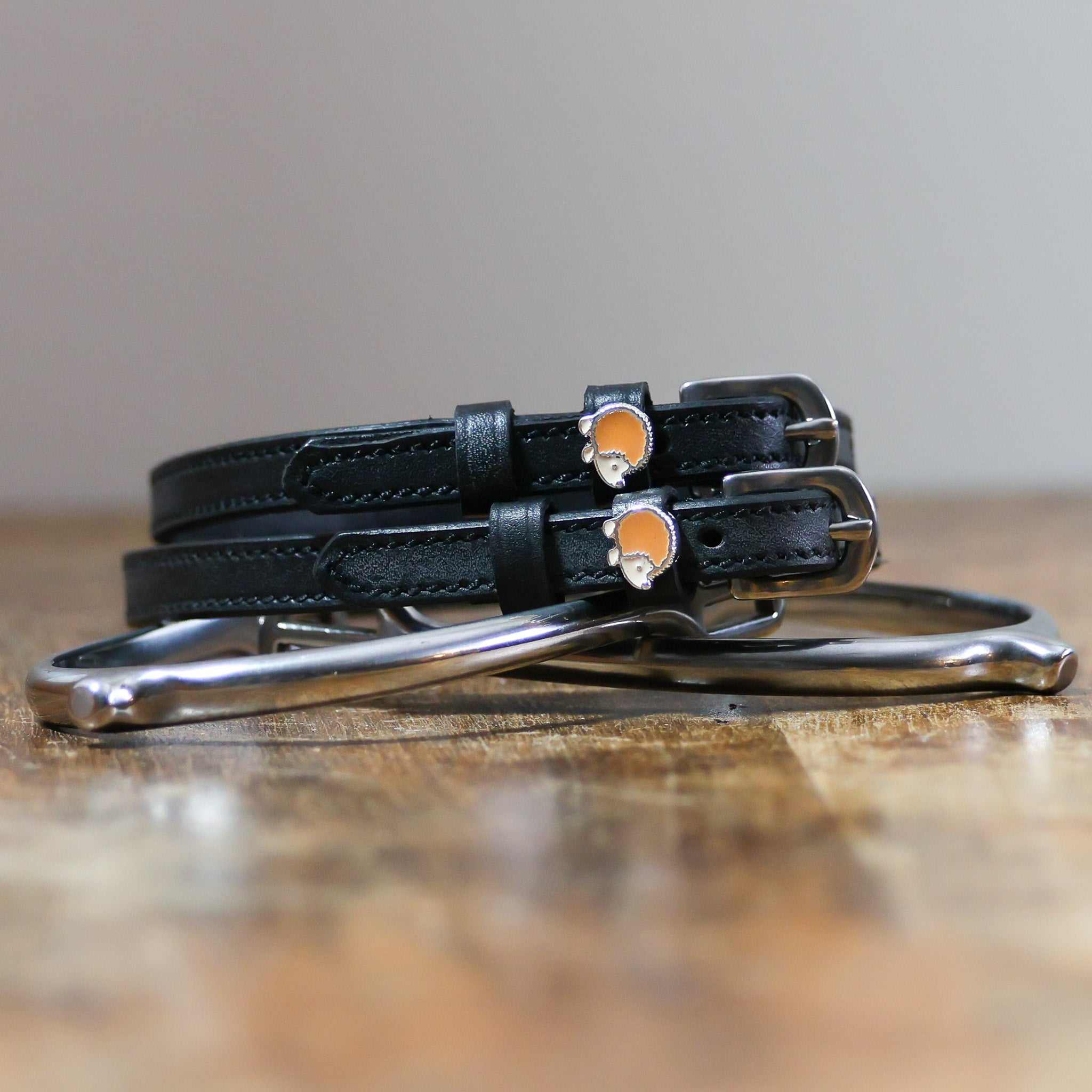 Black leather hedgehog spur straps with stainless steel buckles and custom charms on wooden surface