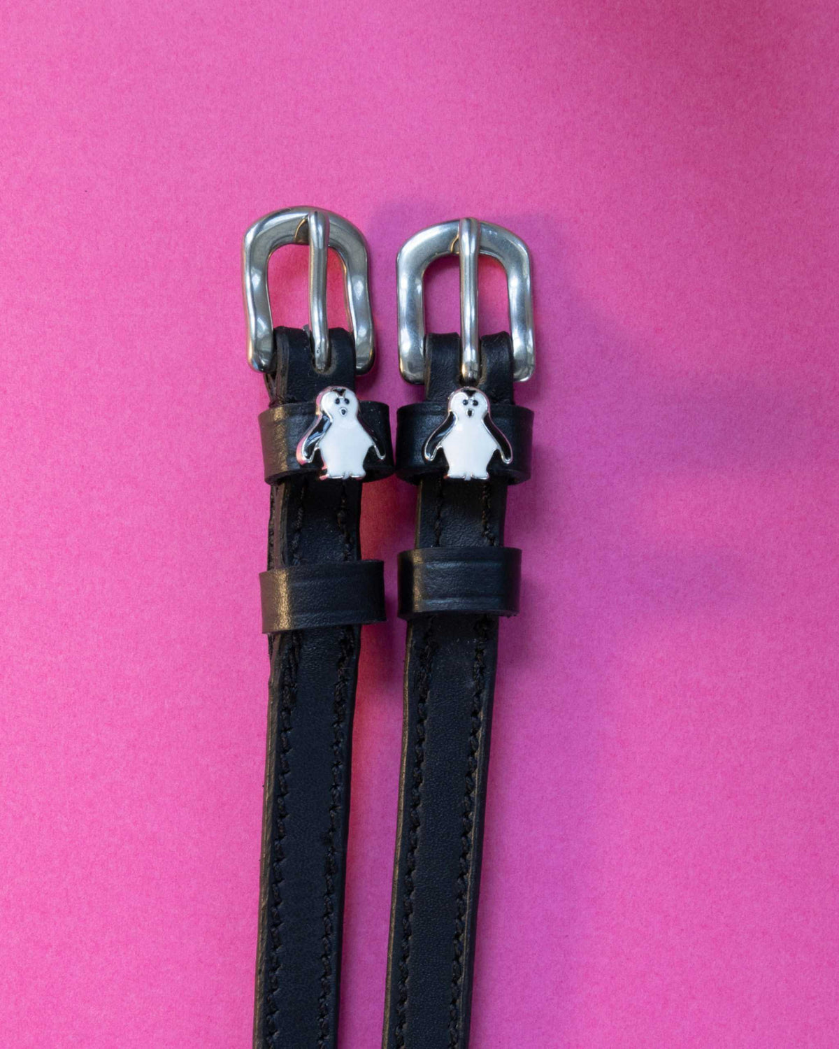 Penguin ManeJane spur straps with the signature Penguin charm, shown on a magenta background, by Dreamers & Schemers
