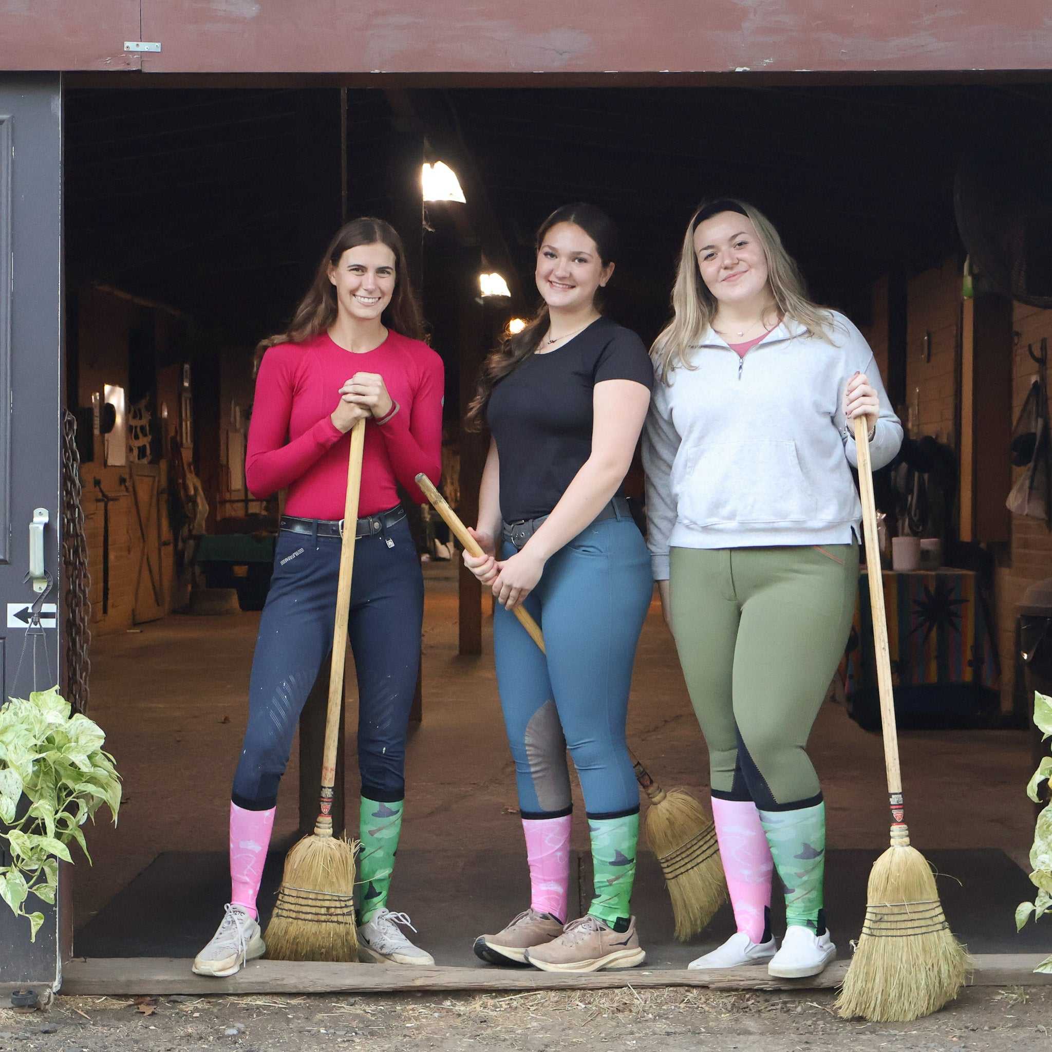 Three women wearing colorful boot socks standing with brooms in a barn entrance