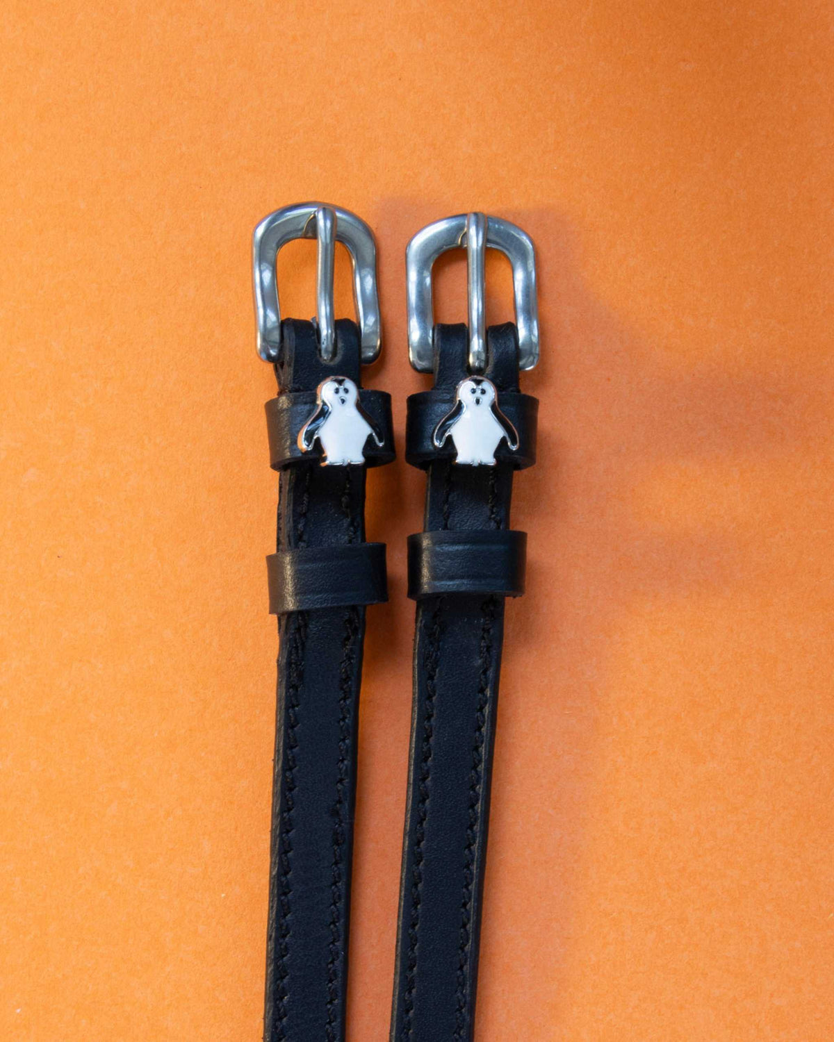 Penguin ManeJane spur straps with the signature Penguin charm, shown on an orange background