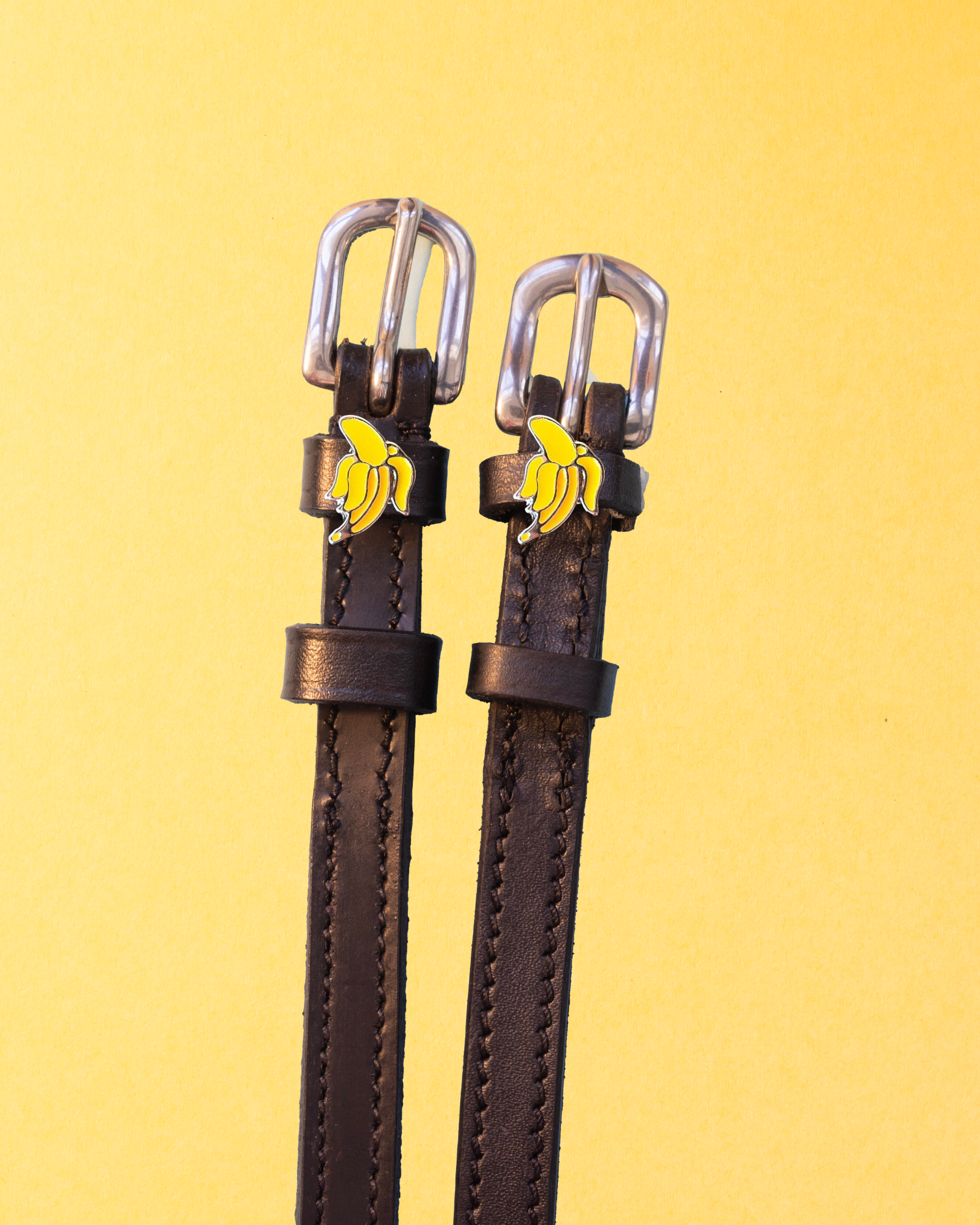 Dark brown leather banana charm spur straps with silver buckles on a yellow background