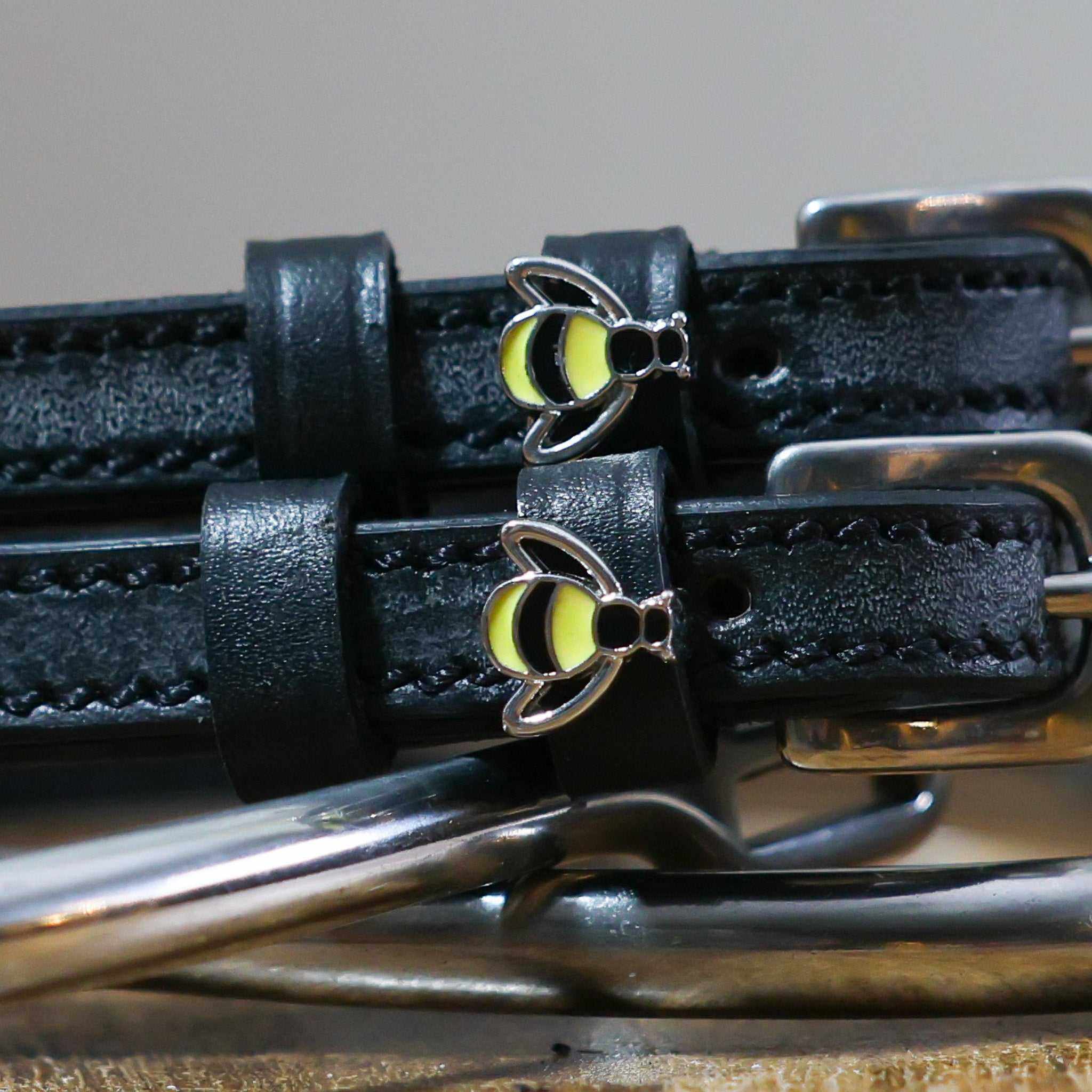 Close-up of silver-and-yellow bee charms on black leather ManeJane spur strap keepers