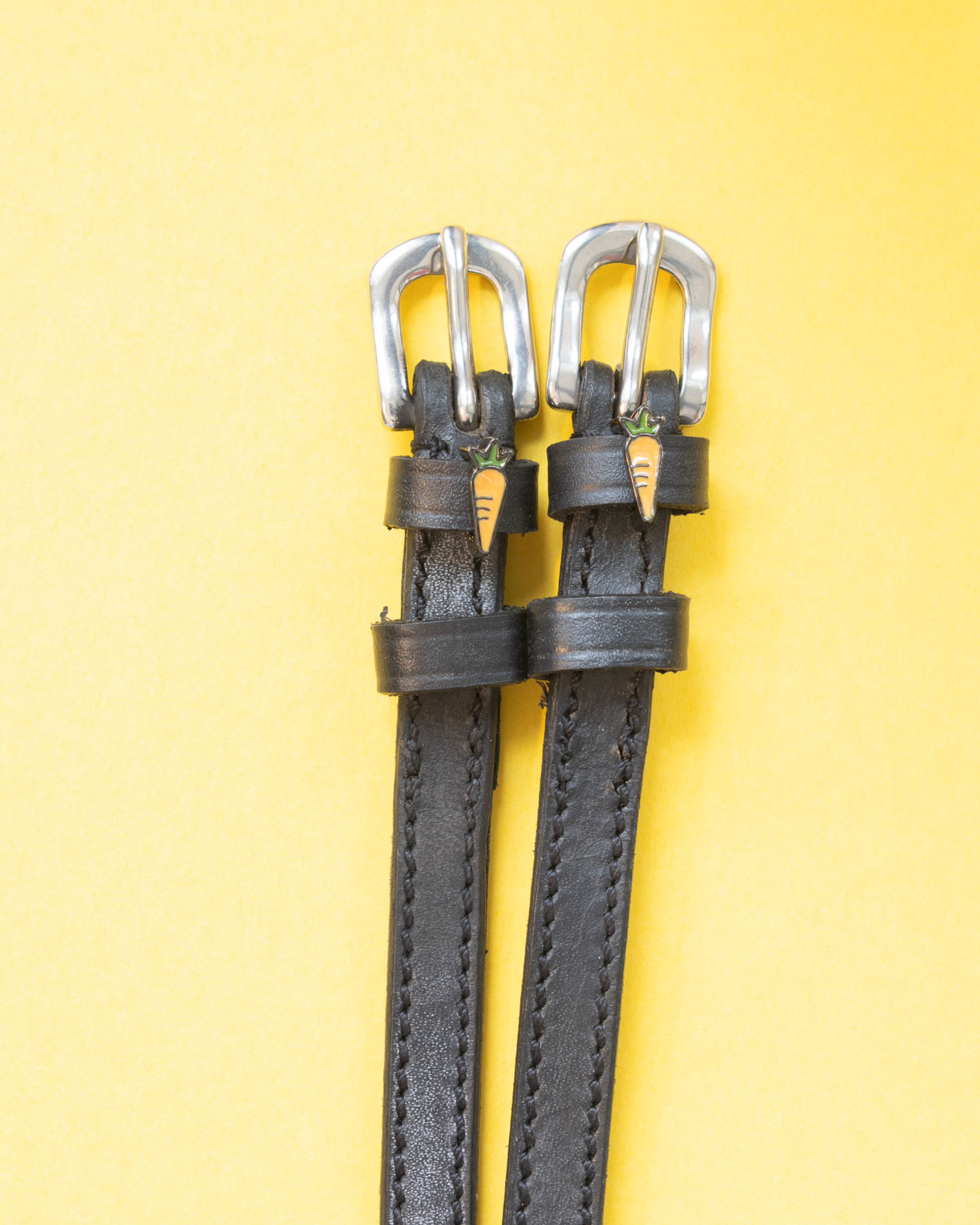 Pair of black leather ManeJane carrot spur straps with silver buckles on a yellow background
