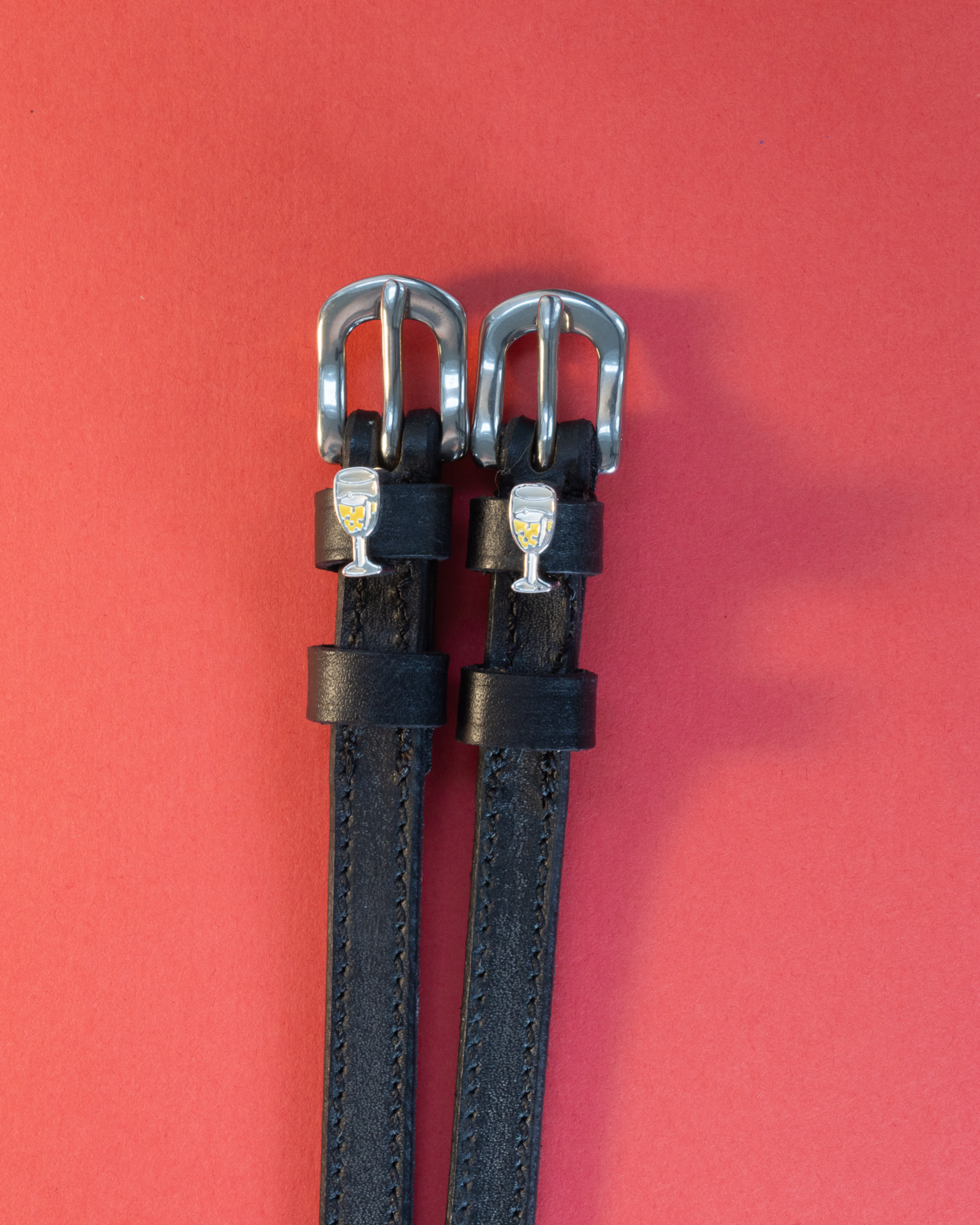 Black leather ManeJane spur straps with champagne-glass charms on a coral background