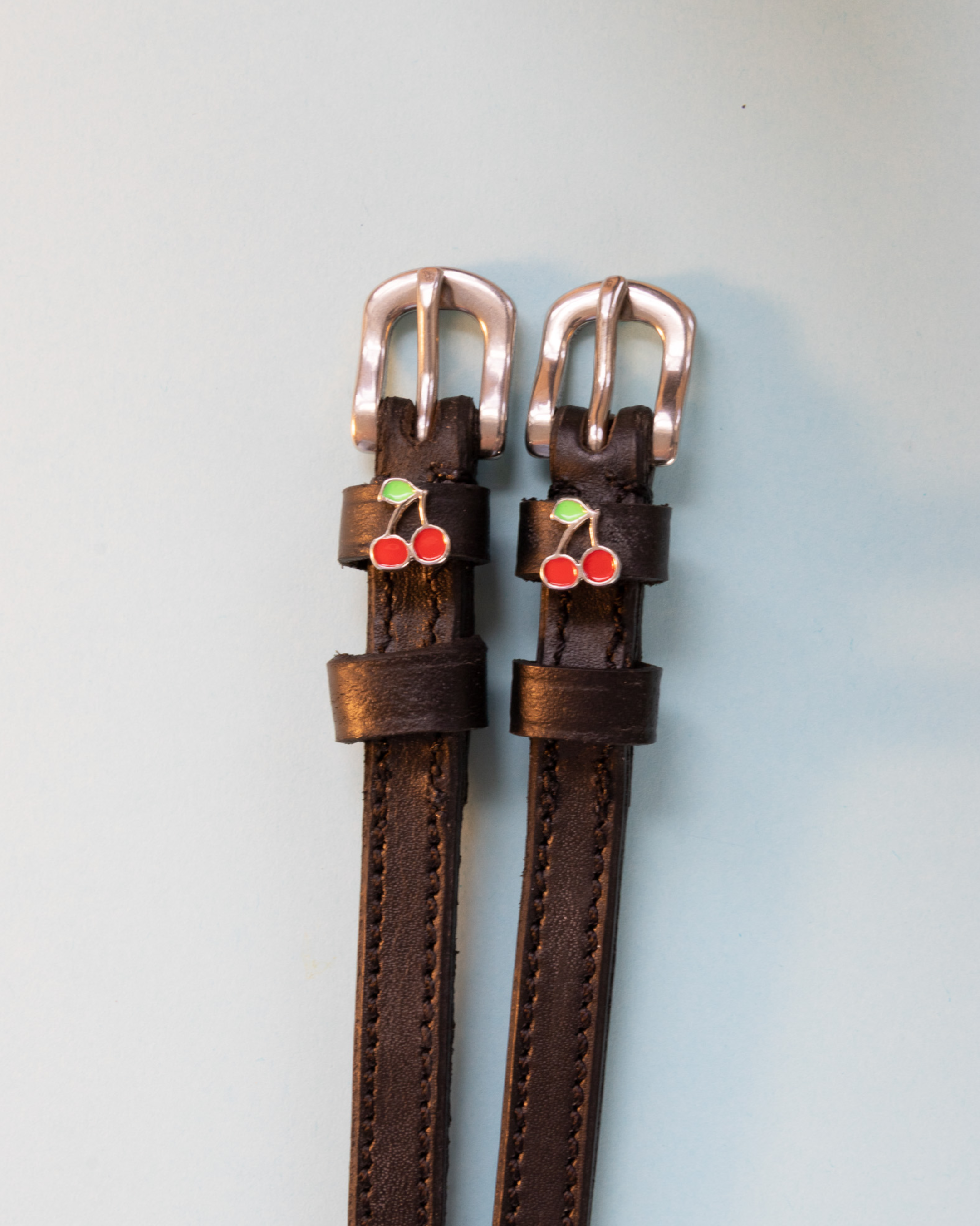Dark brown leather ManeJane cherry spur straps with rose-gold buckles on pale blue