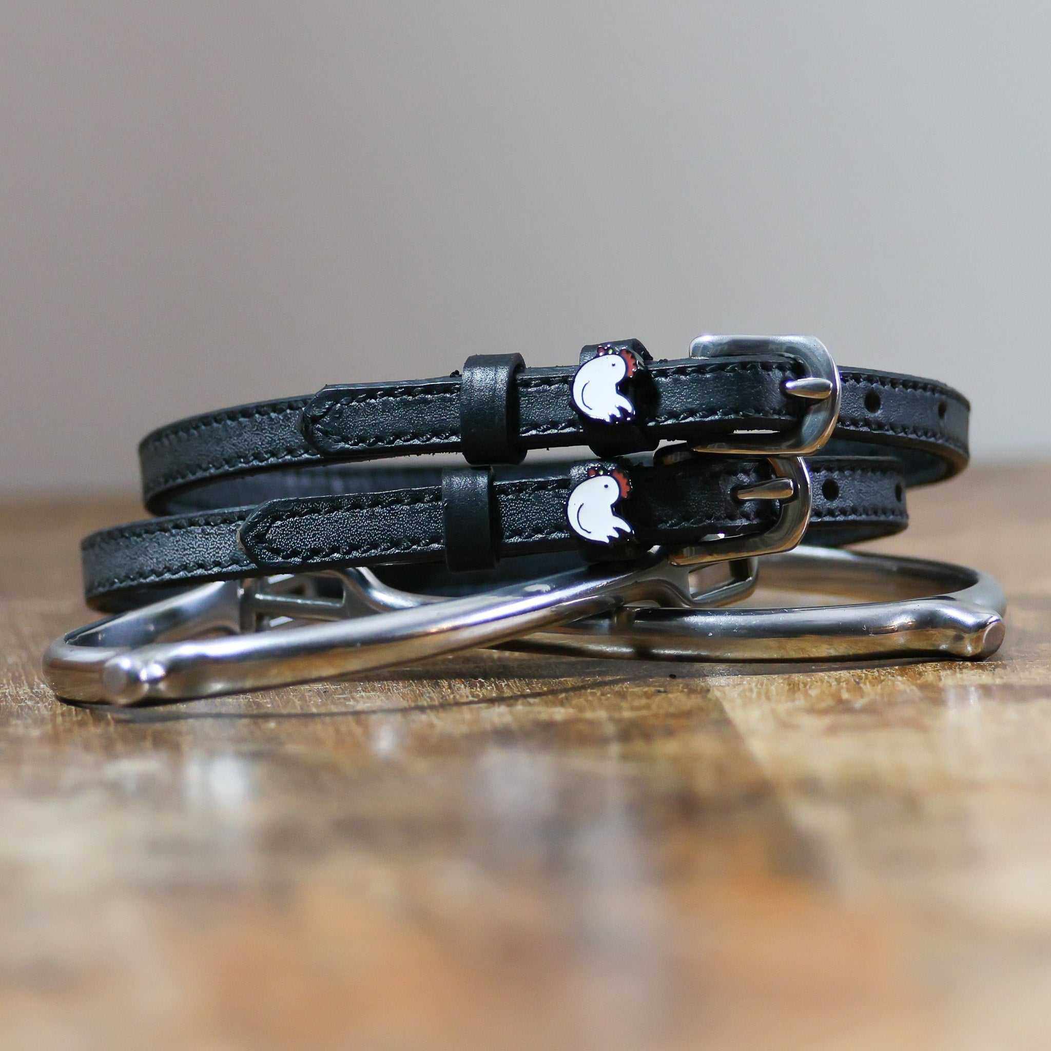 Black leather chicken spur straps coiled beside a silver stirrup on a wooden surface