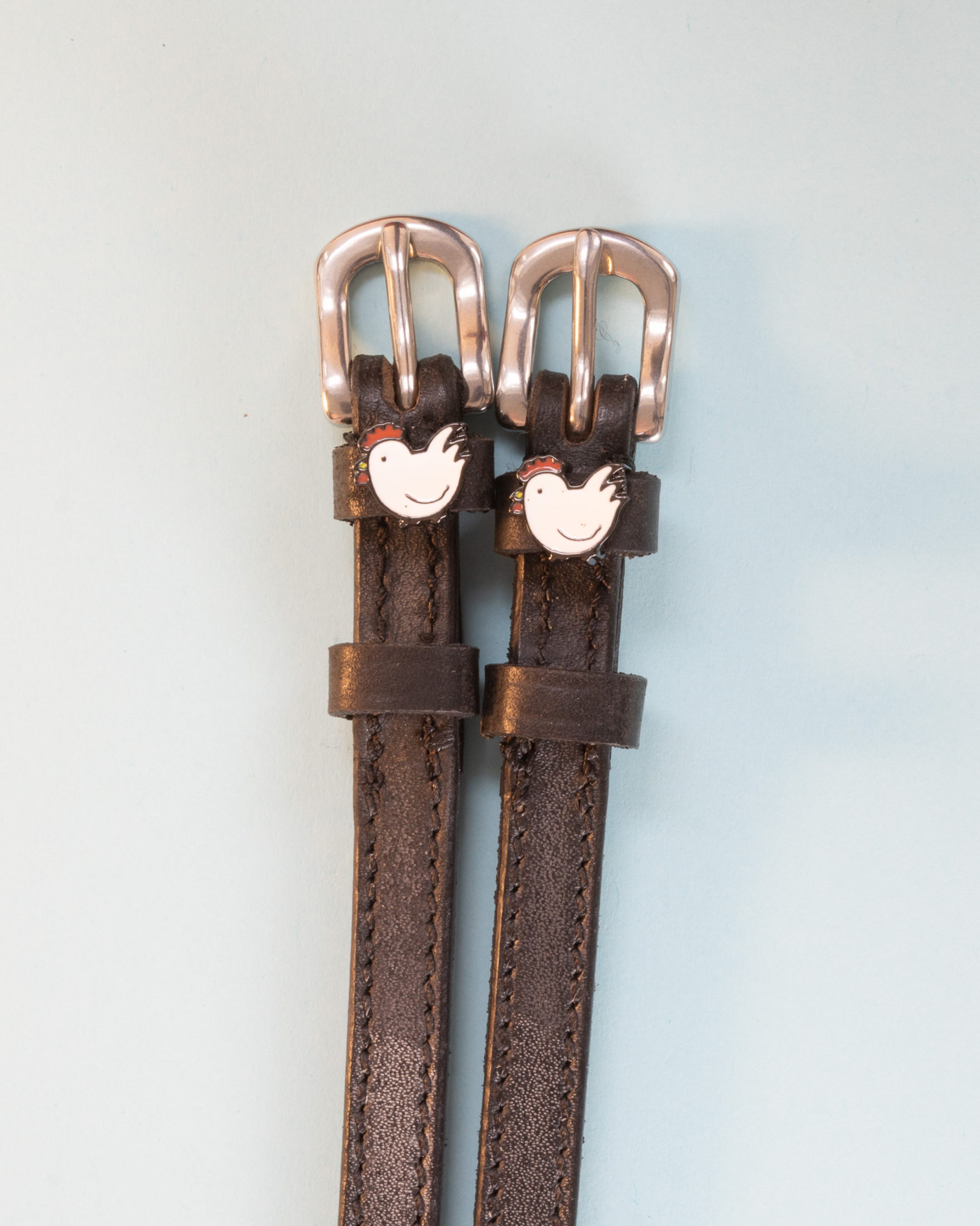 Brown leather ManeJane chicken spur straps with rose-gold buckles on pale blue