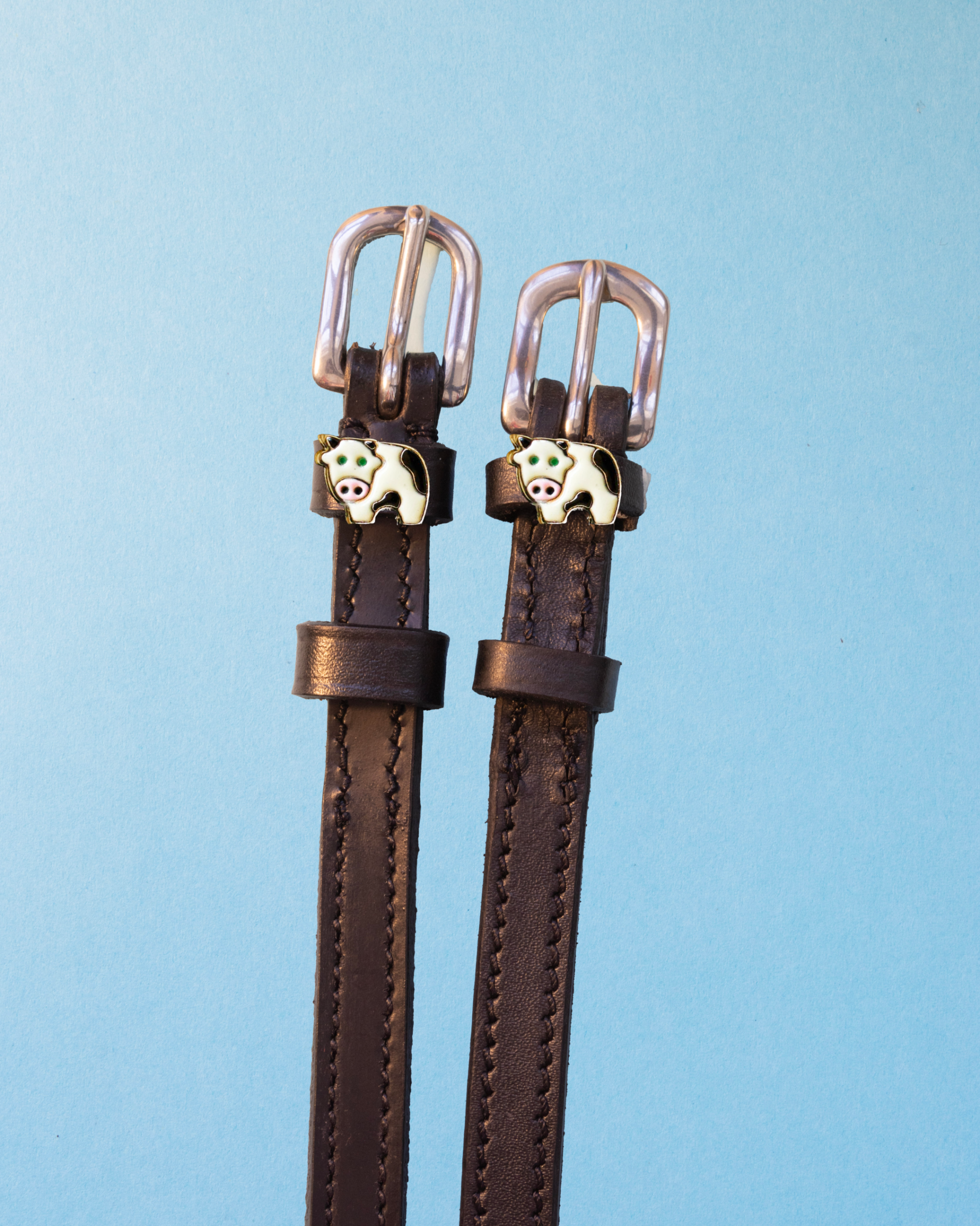 Brown leather cow spur straps with enamel cow charms on a light blue background, ManeJane