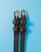 Black leather ManeJane spur straps with round snaffle-bit charms on light blue