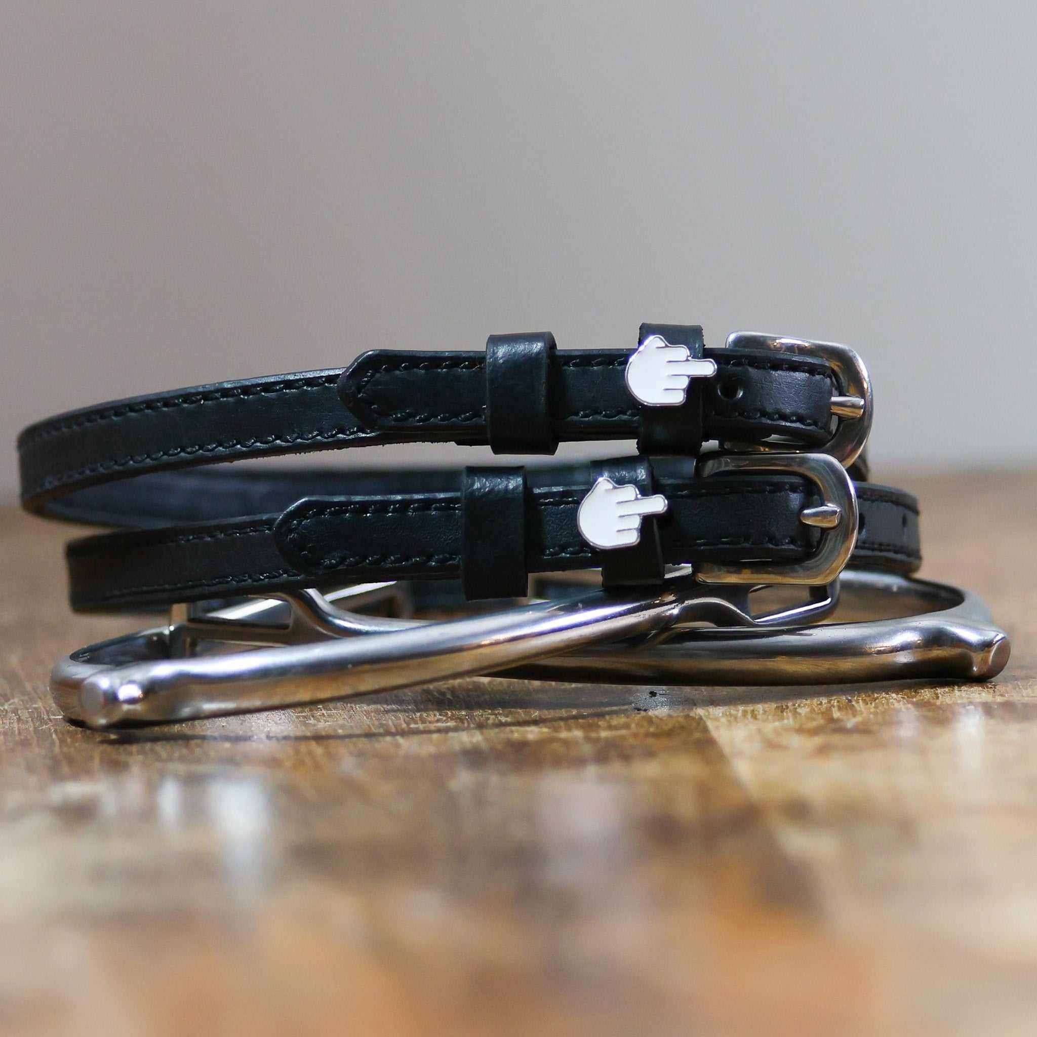 Stacked black Da Bird spur straps with silver buckles on wood beside a stirrup iron, ManeJane