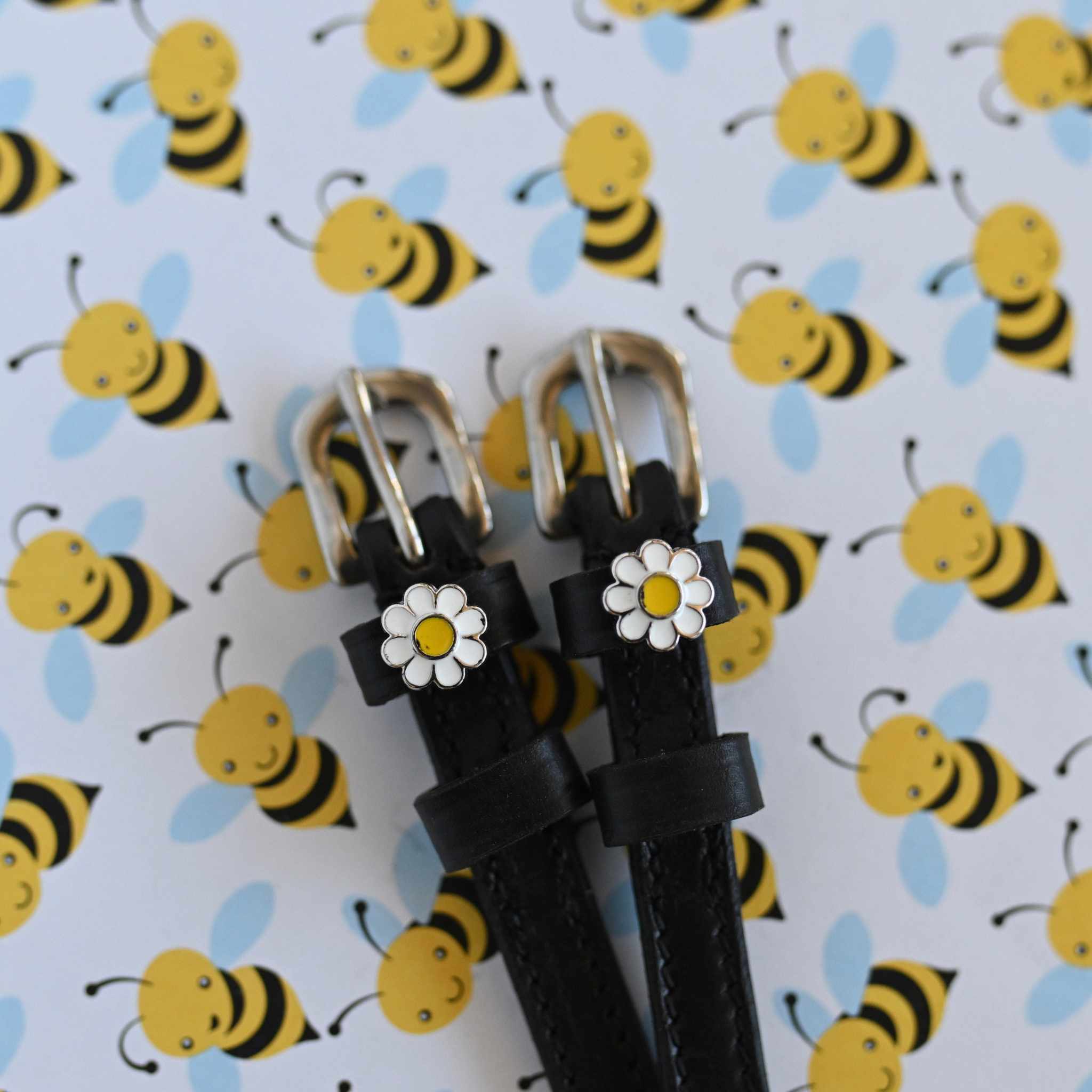 White daisy charm spur straps with silver buckles against a cartoon bee print backdrop