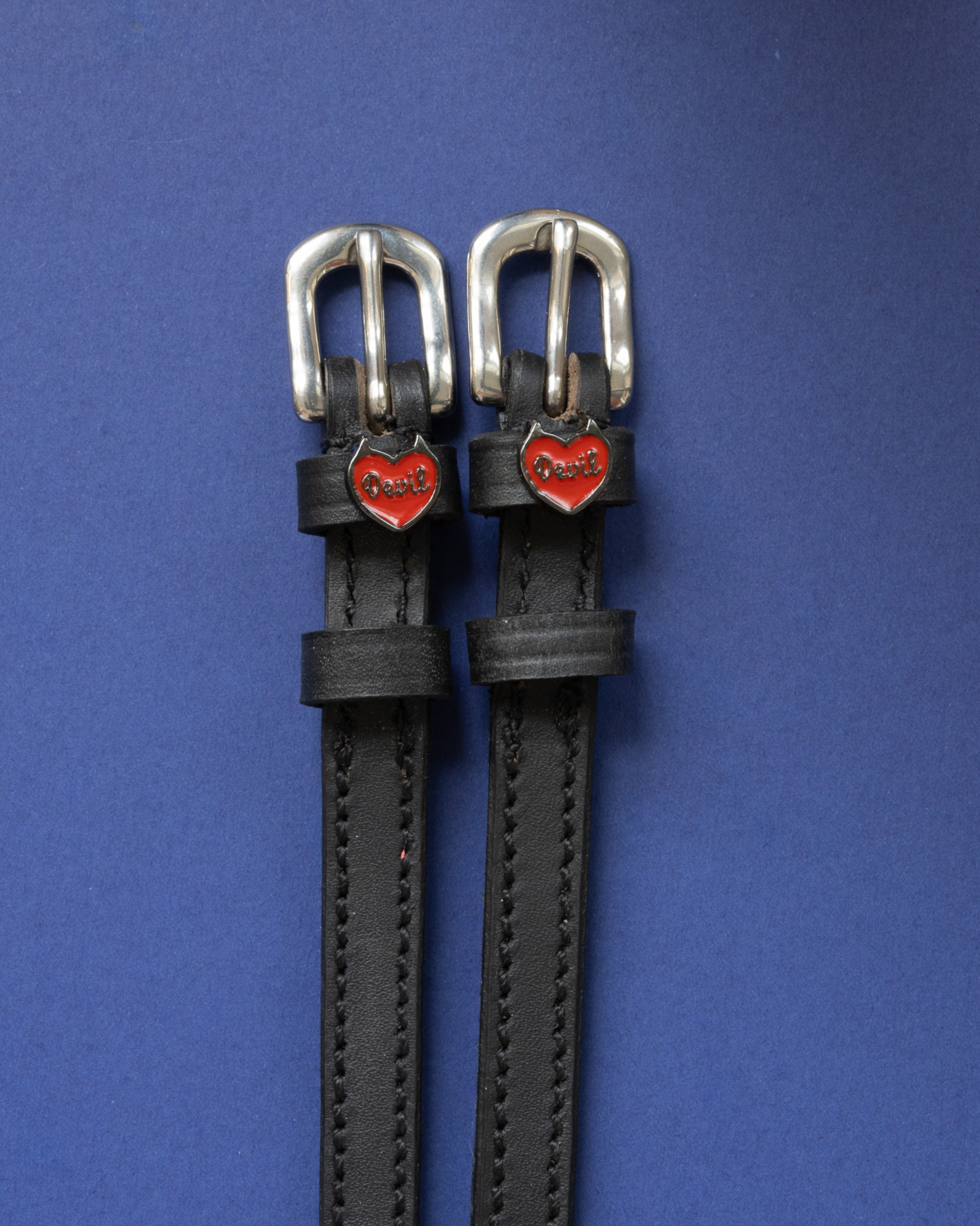 Pair of black leather Devil heart spur straps flat-laid on a royal blue background