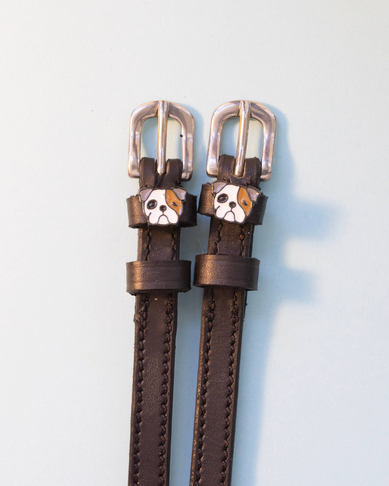 Dark brown leather ManeJane spur straps with bulldog head charms on a pale background